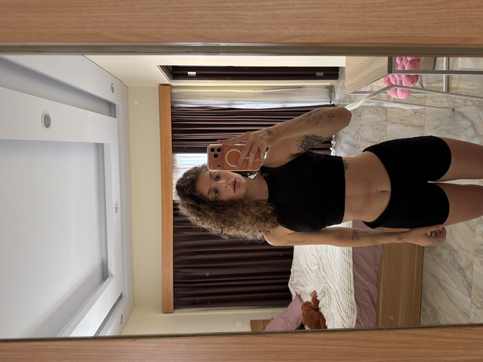A young woman with curly brown hair takes a mirror selfie in what appears to be a bedroom. She is wearing a black crop top and black shorts. Tattoos are visible on both of her arms, and light red marks, possibly from clothing, are present on her abdomen. She holds a smartphone with an orange transparent case, prominently displaying a circular MagSafe ring with a symbol inside it.
The reflection reveals a room with a bed featuring a white knitted blanket and a purple sheet, alongside a wooden headboard. A small brown stuffed animal is visible on the bed. Behind her, a window with dark brown blinds is partially visible, allowing some natural light into the room, suggesting it is daytime. The room's ceiling features recessed spotlights. The floor is covered with light-colored, patterned tiles, contributing to a modern aesthetic. To the right, a white metal stand with pink fluffy slippers is visible. The mirror itself has a light wooden frame. The tiled floor and dark blinds suggest a warm climate, consistent with the specified location of North Kuta, Indonesia. No discernible text is visible apart from the symbol on the phone case.