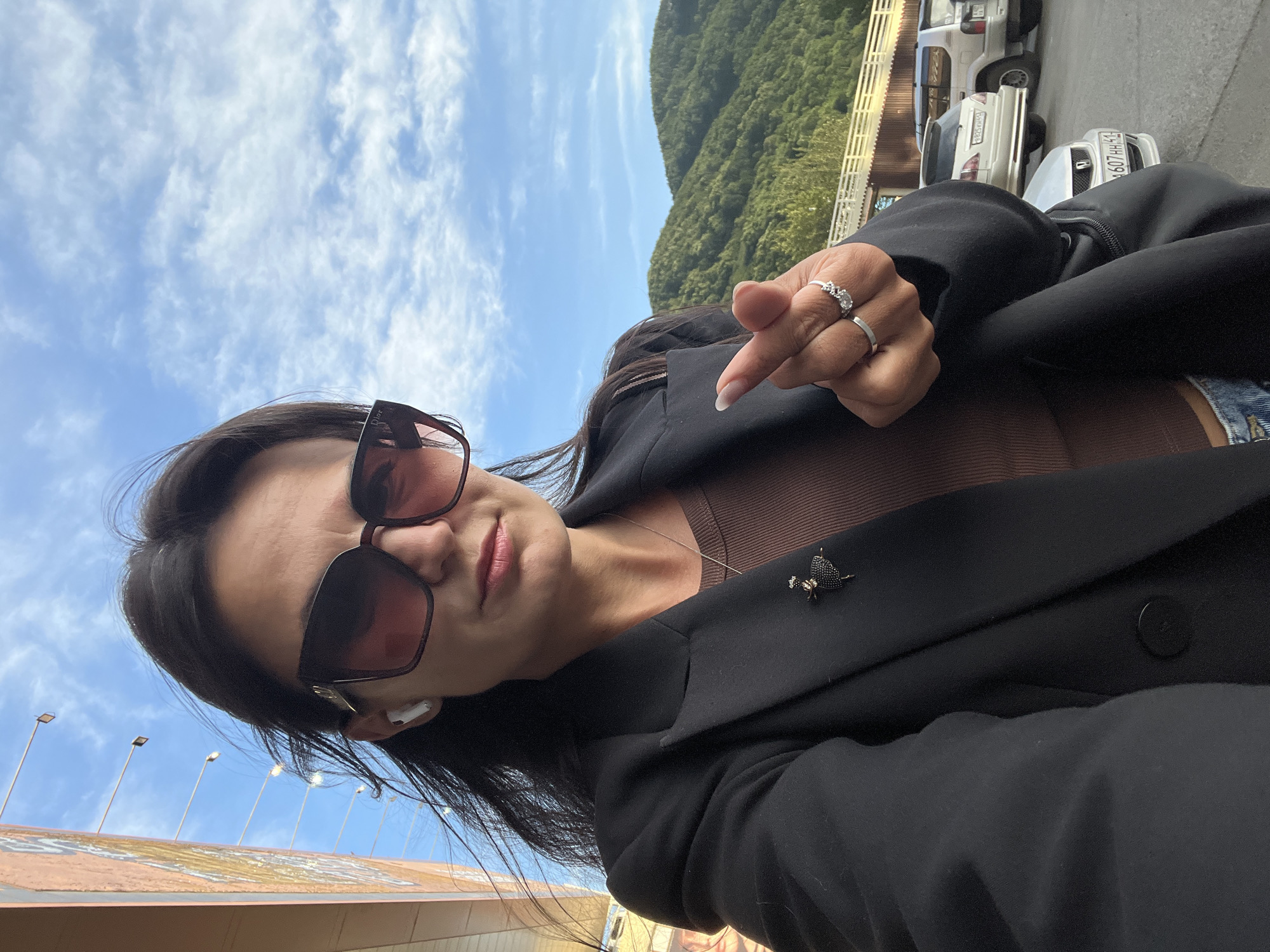A woman with long, dark hair is taking a selfie outdoors. She is wearing large, dark sunglasses, a brown top, and a black blazer adorned with a black brooch in the shape of a dancer. She has a white wireless earbud in her left ear and is making a finger heart gesture with her right hand, which is adorned with two rings. The background features a bright blue sky with scattered white clouds, a light-colored building on the left, and a mountain range covered in green trees on the right. Several cars are parked in the background. The setting appears to be an outdoor area, potentially a parking lot or roadside, near a city. It is unlikely to be Petropavlovsk-Kamchatsky, Russia, due to the absence of distinctive geographical features or urban elements commonly associated with the city.
