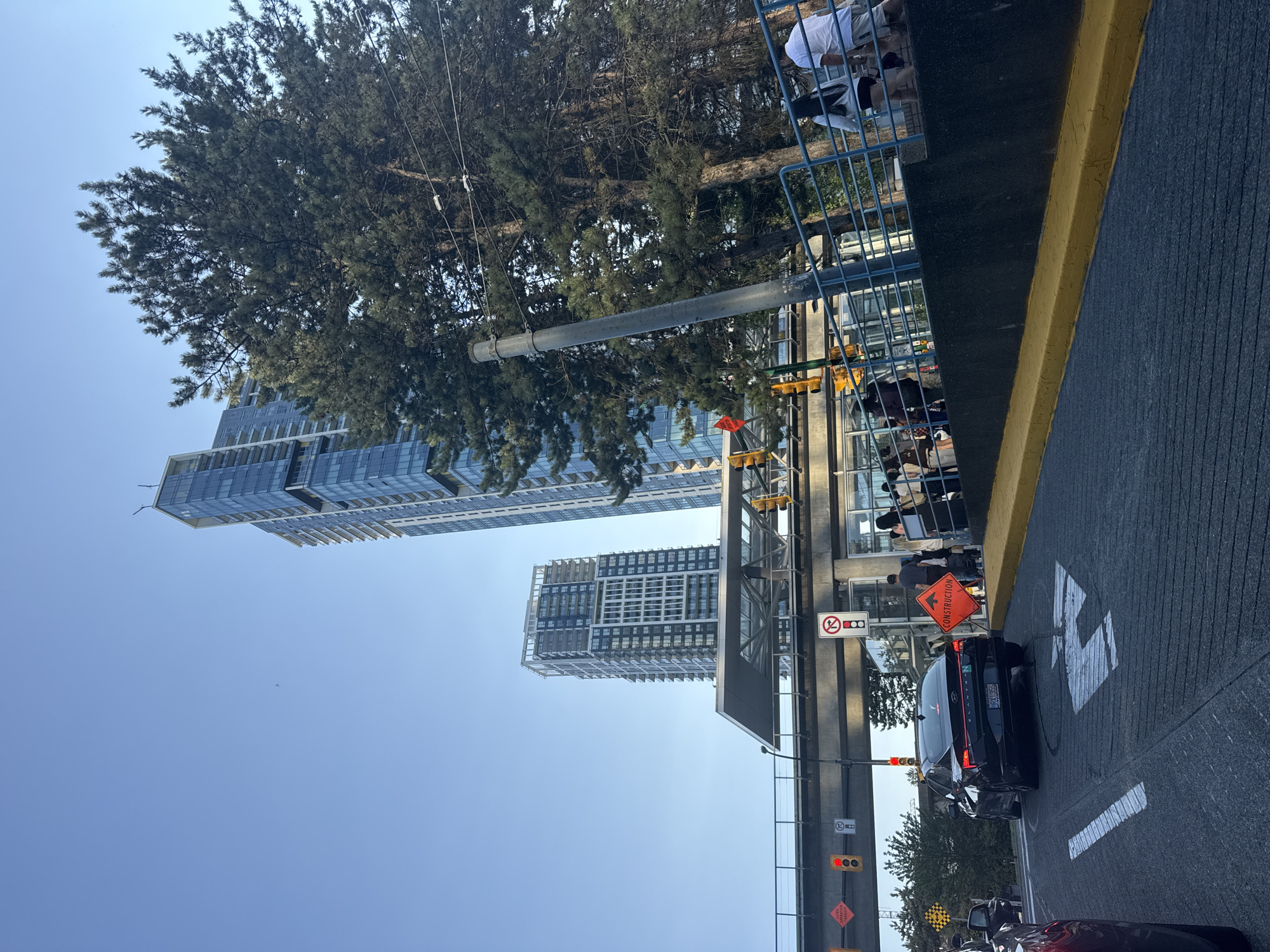 The image shows a street scene in Burnaby, Canada, with two tall, modern apartment buildings in the background under a clear blue sky. A pedestrian overpass, likely part of a transit system, spans the street in the middle ground. Several traffic lights are visible, with one showing red. An orange diamond-shaped sign with the word "CONSTRUCTION" is present on the right side of the road.
In the foreground, a black Hyundai Elantra is turning left. There are lane markings on the asphalt indicating a left turn. People are visible on the pedestrian overpass, some walking and some standing. A group of people are standing on the right side of the overpass, near a blue railing. A tall metal pole, possibly a light pole or utility pole, stands near the railing. Trees with dense green foliage are situated to the right of the buildings and behind the overpass. The overall impression is of an urban environment with a transit hub.
