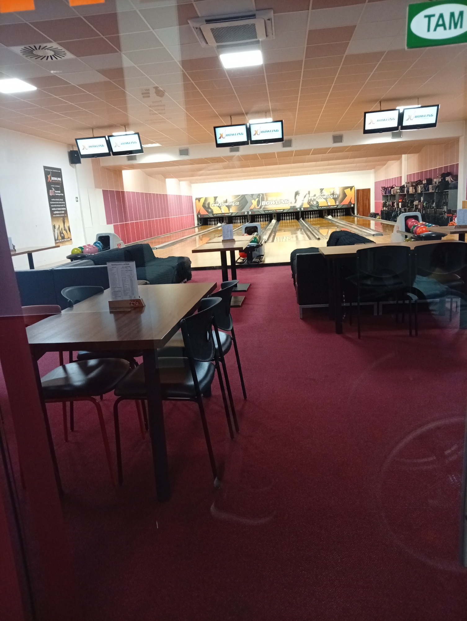 The image depicts the interior of a bowling alley. In the foreground, a wooden table is set with chairs, suggesting a seating area for patrons. A menu is placed on a wooden stand on the table. Further back, a dark sofa is visible, and beyond that, several bowling lanes stretch into the distance. Bowling balls of various colors are arranged in a rack. Above the lanes, screens display the word "BOWLING" and some logos. In the far background, there are racks of bowling shoes and bags. A sign with the word "TAM" in green letters is visible on the ceiling in the upper right corner. The scene appears to be set during the day, with artificial lighting illuminating the space. The overall atmosphere is that of a recreational facility. The word "BOWLING" is prominent on multiple screens, and "xbowling.cz" is visible on a banner above the lanes. The sign "TAM" might indicate a brand or a section of the venue.