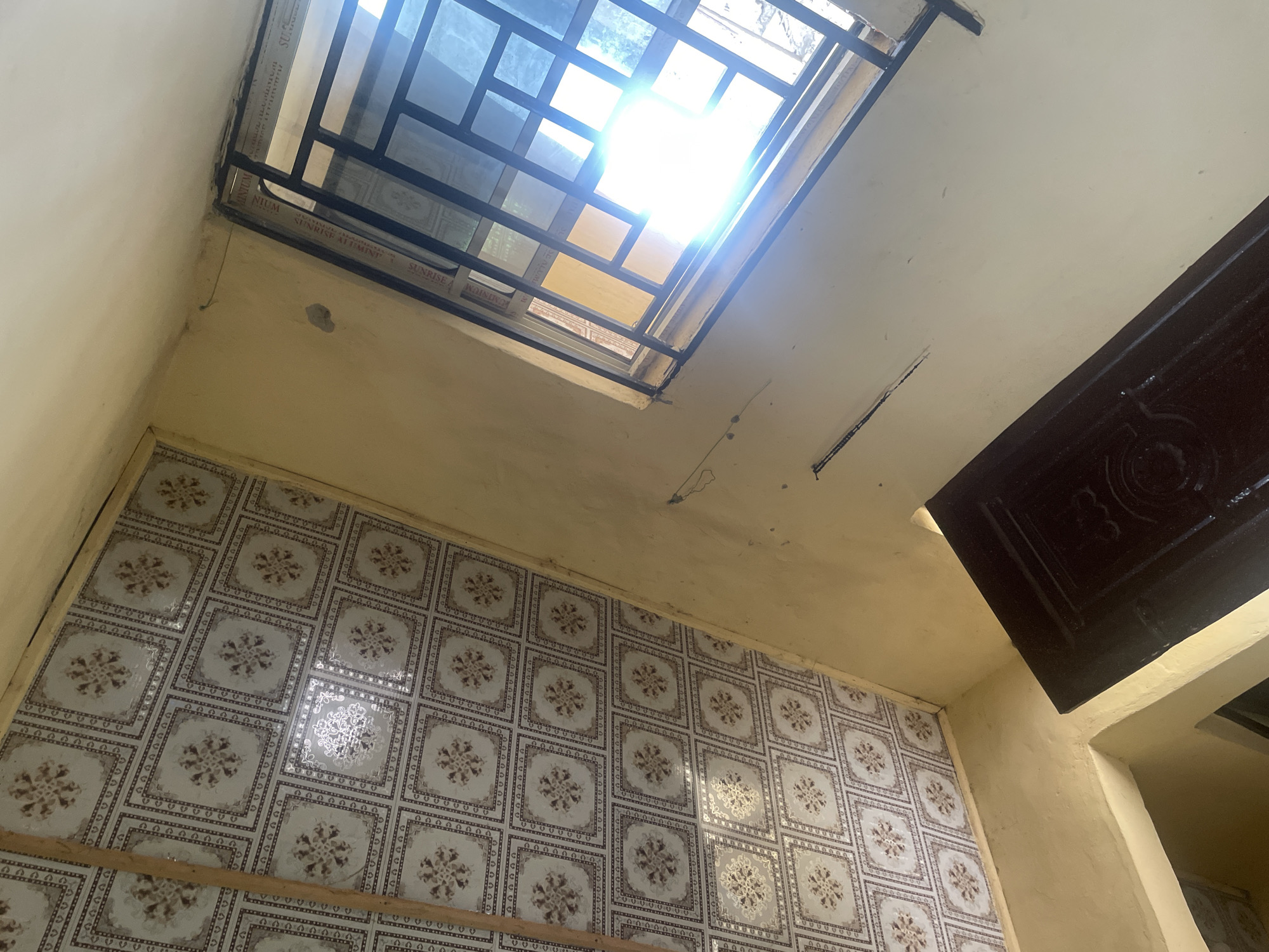 An interior shot looking up at a ceiling, a window, and a door in a building in Jalingo, Nigeria. The ceiling is covered in patterned tiles with a floral motif. A wooden beam is visible along the edge of the ceiling. To the right is a window with a metal grille. Bright sunlight is streaming through the window, causing a lens flare effect. The walls are painted a light yellow color. A dark brown door is visible in the lower right portion of the image. The overall impression is of a humble dwelling or a functional room.