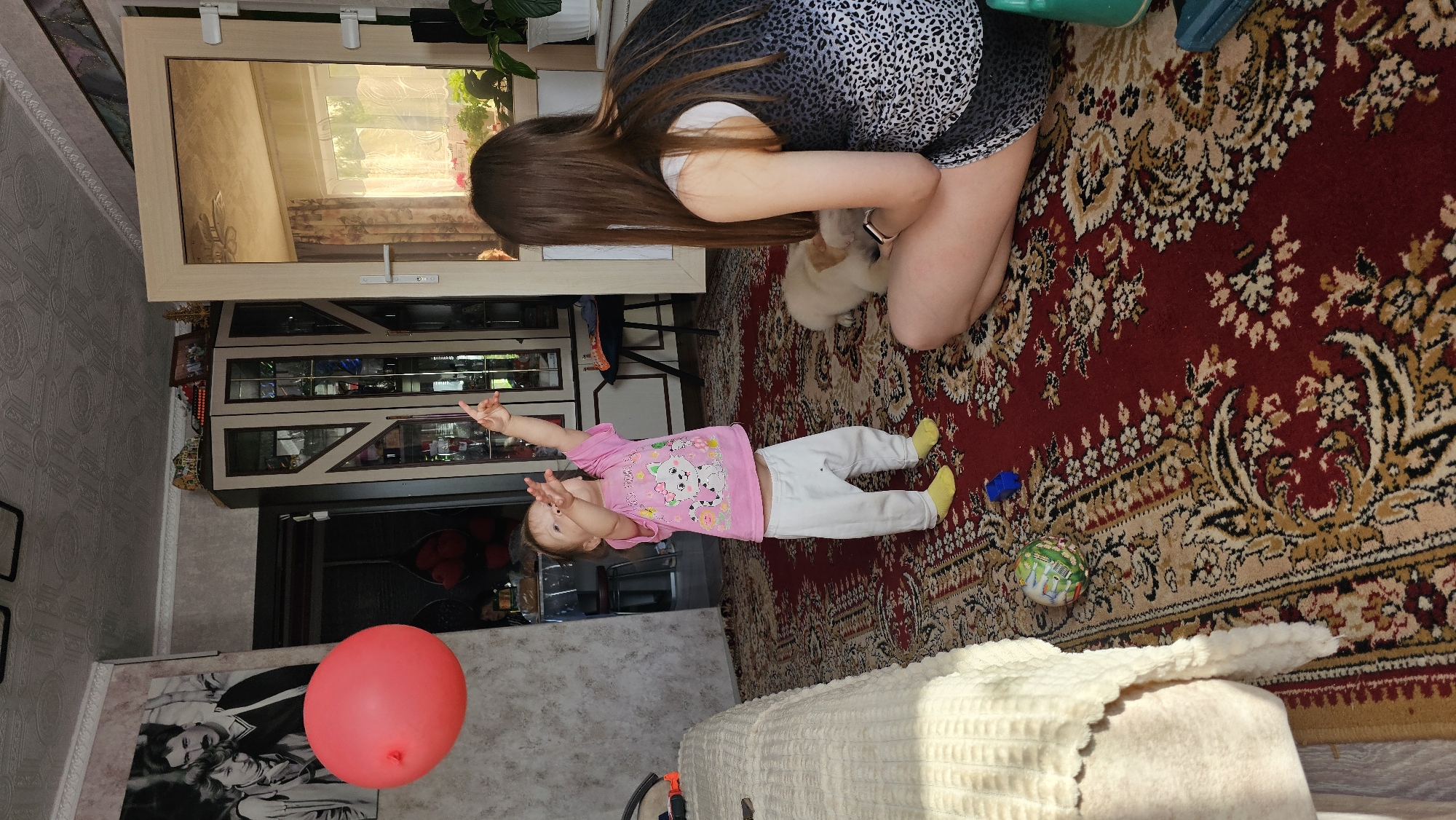 A scene captures a young child lying on an ornate rug, wearing a pink shirt and light pants with yellow socks, pointing upward with both arms raised. An adult woman, seen from the shoulders down, is kneeling beside a small, fluffy white dog. The rug is a vibrant red with gold and black patterns, taking up a significant portion of the floor space. In the background, a glass-fronted cabinet displays various items, and a red balloon floats near a couch. A framed picture hangs on the wall. The location is a domestic interior.