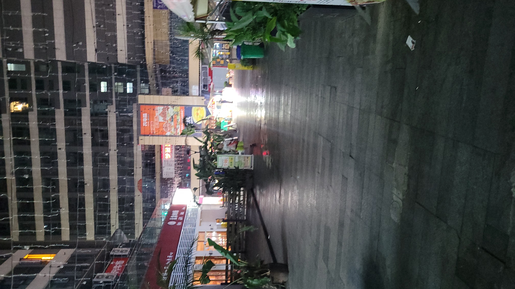 The image depicts an outdoor pedestrian area at night in Jiulongpo District, China. A tall building with multiple floors and vertical stripes dominates the left side of the frame. Various signs and advertisements are visible on the building's facade. The pedestrian area, constructed of pavement, runs horizontally across the image. Several plants and street furniture are located along the walkway. Bright lights emanate from storefronts further down the pedestrian path, creating a strong light-to-dark gradient in the image. No people are clearly visible.