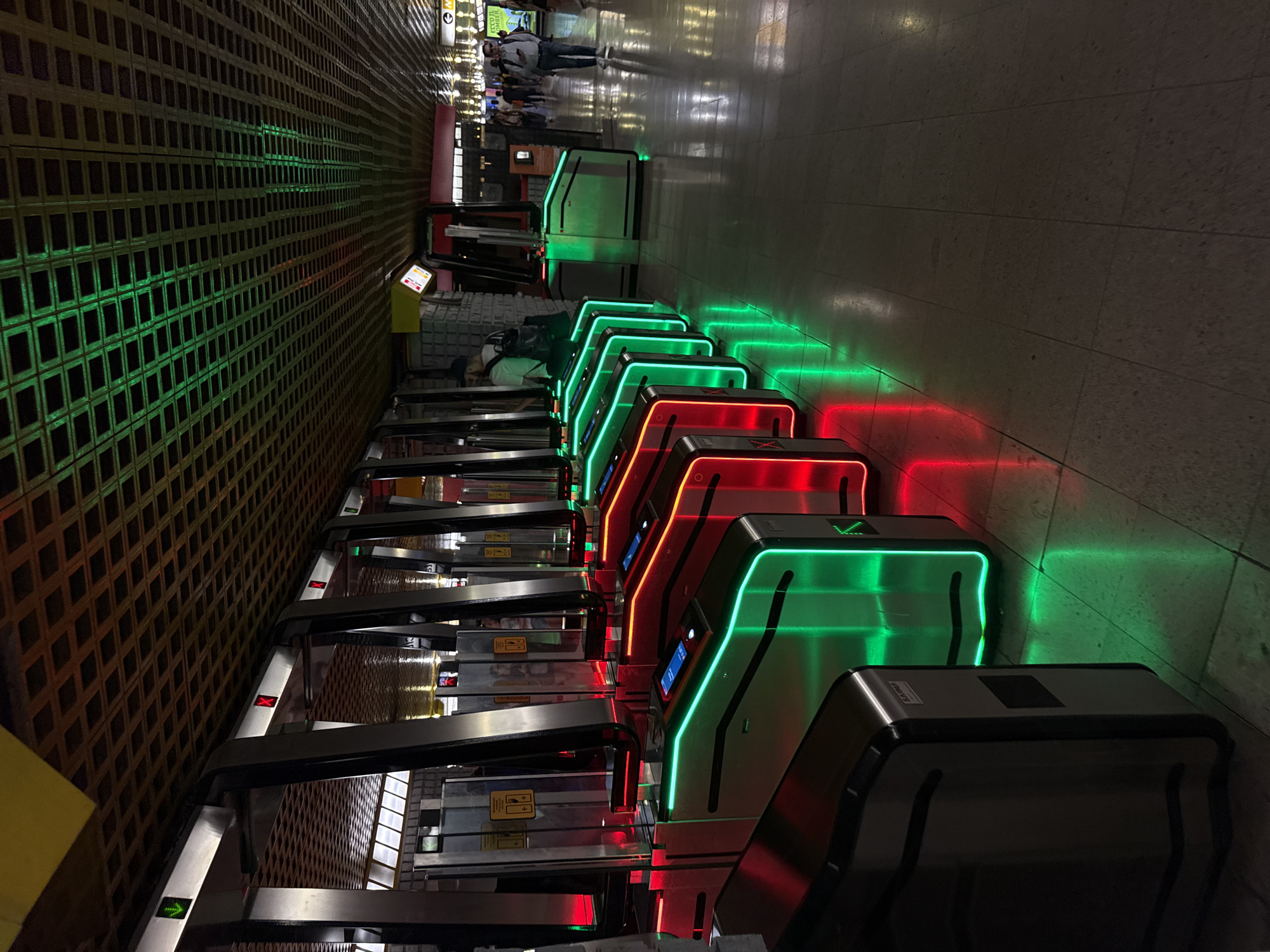 The image captures a row of illuminated turnstiles at a Milan Metro station, likely in a contemporary setting with a patterned ceiling. The turnstiles are emitting vibrant green and red lights, with some displaying digital screens. The floor is tiled and reflects the colorful lights. In the background, several people are visible, some walking away from the turnstiles and others in the distance, suggesting the station is active. The overall impression is a modern, well-lit public transportation hub.