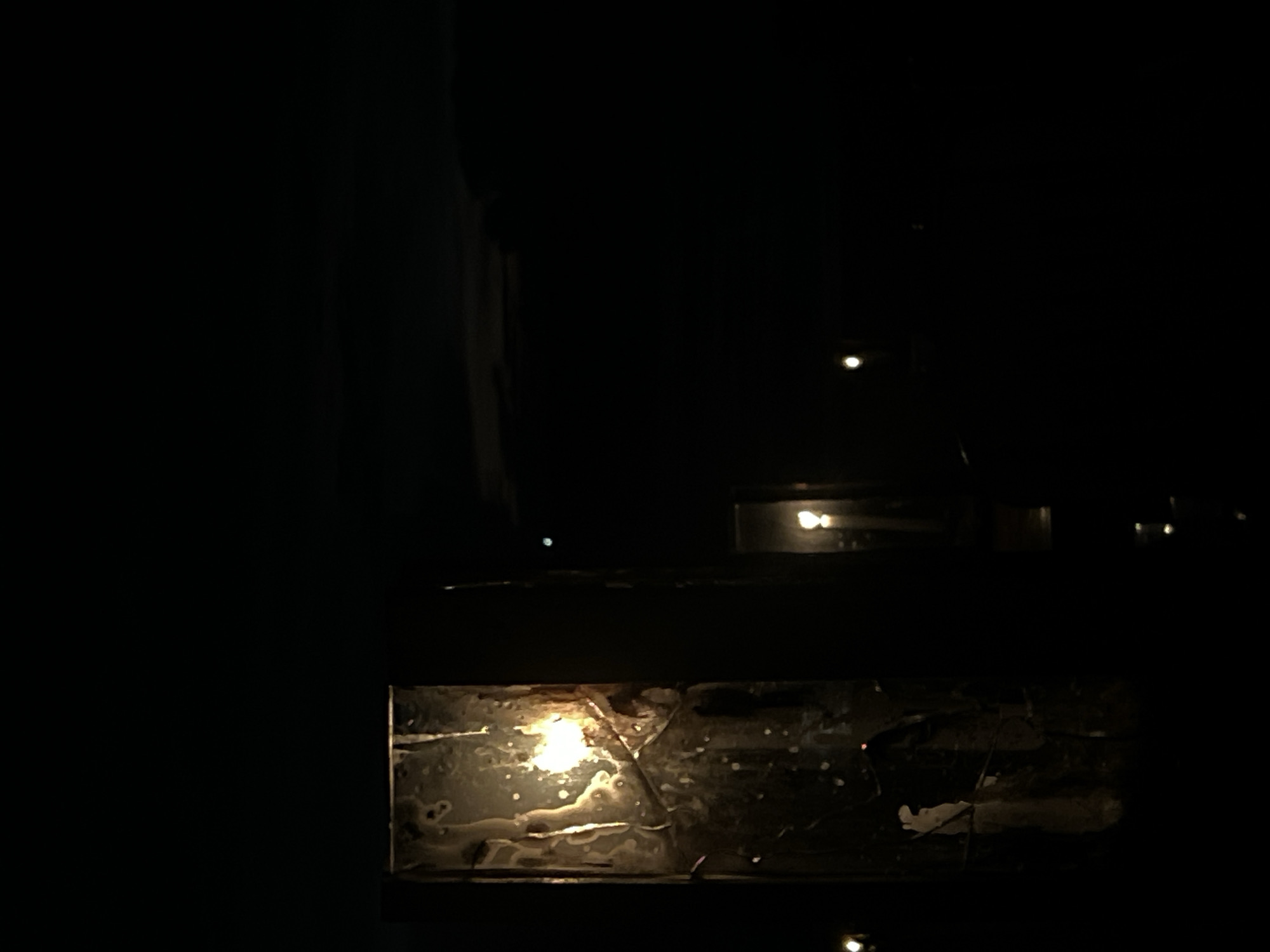 The image is a low-light shot, possibly taken at night in an urban area. A window with a damaged or textured surface is prominent in the foreground. A light source, possibly a candle or small lamp, is visible through the window's imperfections. In the background, there are faint lights indicating the presence of buildings or streetlights. The overall scene conveys a sense of darkness and limited visibility.
