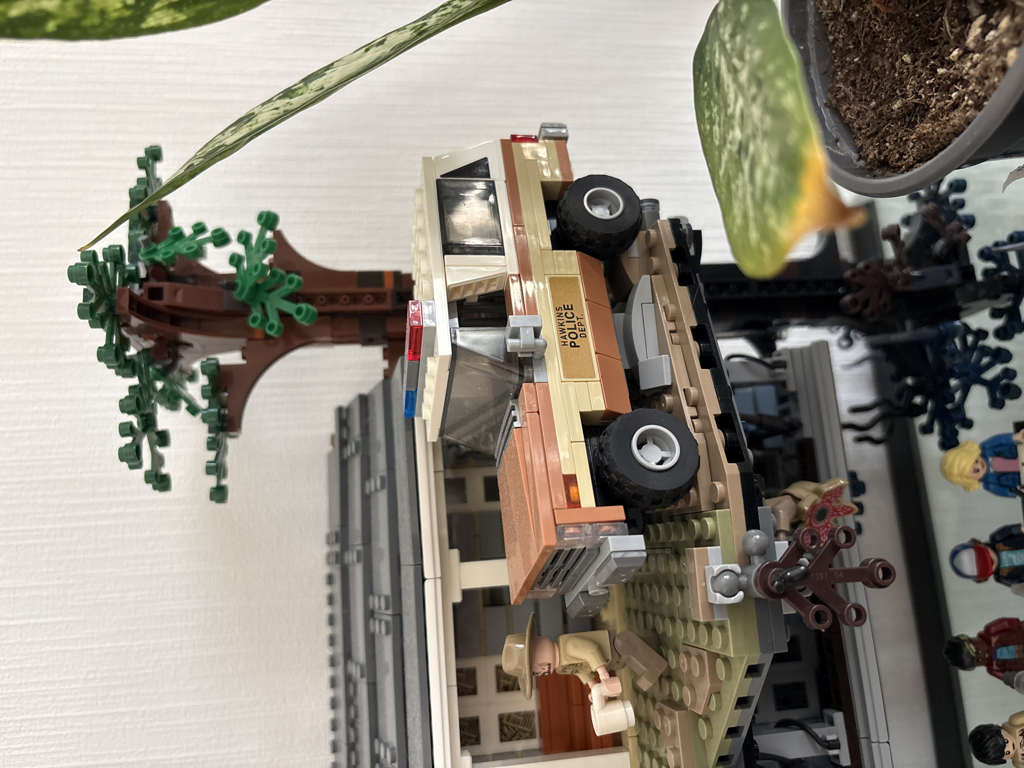 The image depicts a Lego set resembling a scene from the TV show Stranger Things. It includes a replica of the Byers' house and a Hawkins Police Department vehicle. A Lego minifigure in a beige outfit, possibly representing Jim Hopper, is visible near the front door. Additional Lego minifigures are present, seemingly representing other characters from the show, including Demogorgon. A tree and potted plant are visible, possibly for decorative purposes. The scene is set on a white surface.
