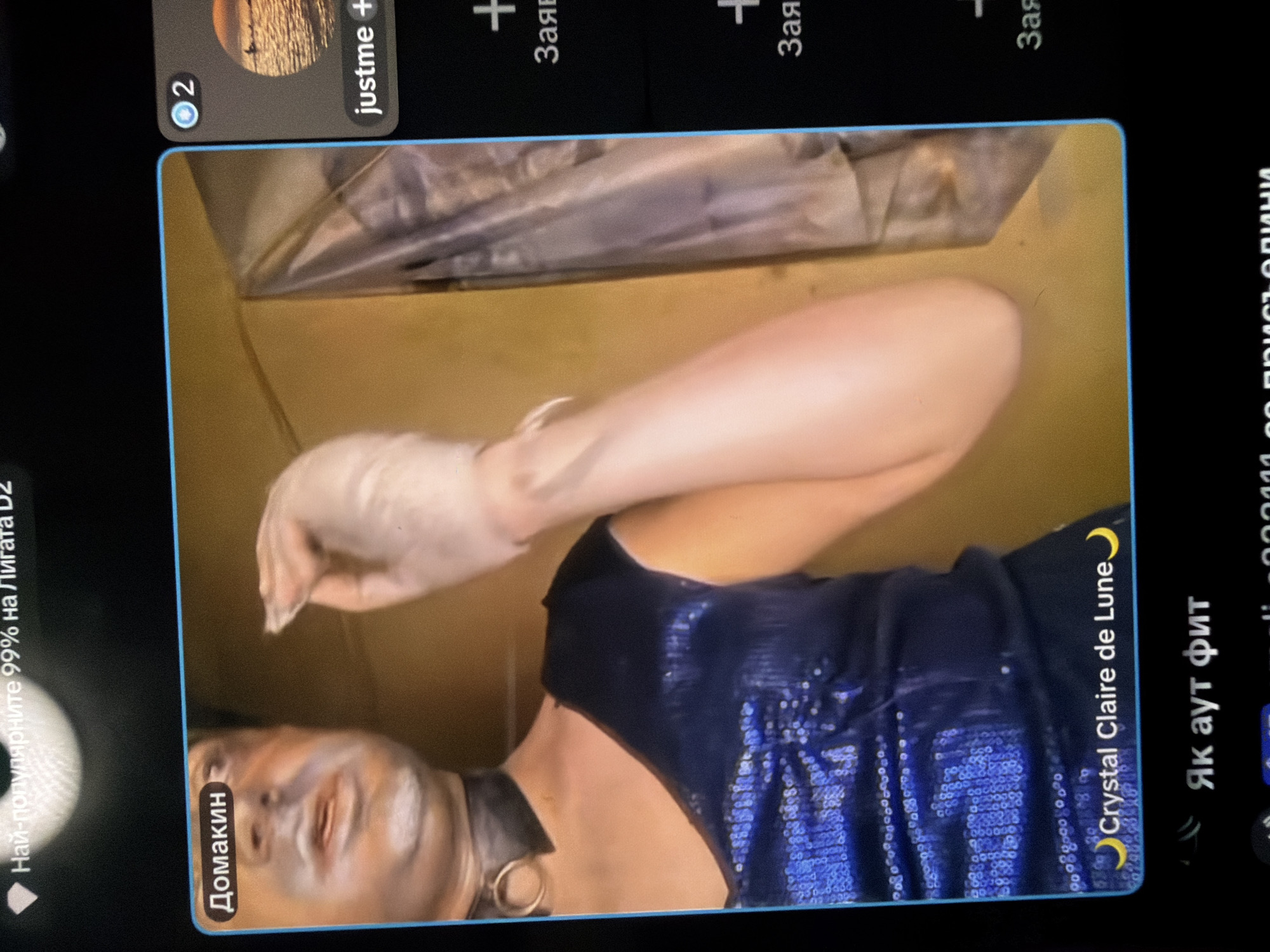 The image is a screenshot of a digital interface displaying a stylized portrait of a person. The main subject, appearing to be female or presenting as female, is lying down against a golden-yellow upholstered surface, possibly a sofa. They are wearing a dark, possibly navy blue, sequined top. Their face and neck are covered in a silvery-gray or pale metallic makeup, complemented by a dark choker with a metallic ring. One arm, also covered in the same silvery-gray substance or a white glove/sleeve, is bent upwards. Behind the person's head, there's a crumpled piece of shiny, metallic-looking fabric acting as a backdrop or prop. The overall aesthetic suggests a theatrical performance, photoshoot, or artistic expression.
The image is rotated 90 degrees counter-clockwise within the screenshot frame. The surrounding digital interface shows various text overlays and elements. Labels visible on the main image include "Домакин" (Host), "Crystal Claire de Lune 🌙", and "Як аут фит" (likely "What an outfit" in Bulgarian, transliterated from English "outfit"). Outside the main frame, text elements include "Най-популярните 99% на Лигата D2" (The most popular 99% in League D2), "justme +", "Заяв" (Request), and a number "2", indicating viewers or interactions. The presence of Cyrillic text in Bulgarian aligns with the specified location context of Sofia, Bulgaria. The lighting appears to be artificial, highlighting the metallic elements of the costume and makeup.