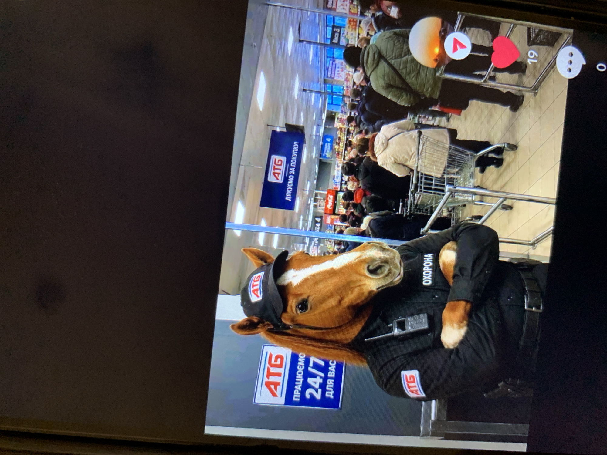 A person wearing a horse mask and an AT G security uniform stands with arms crossed in front of a grocery store in Lymanka, Ukraine. The uniform includes a black shirt with an AT G logo on the left sleeve and the word "ОХОРОНА" (meaning "security" in Ukrainian) on the right breast, a black cap with an AT G logo, and a radio clipped to the shirt. A sign behind the person reads "ATG ПРАЦЮЄМО 24/7 ДЛЯ ВАС" (meaning "ATG We work 24/7 for you"). In the background, shoppers are seen in the aisles of the store, with AT G branded signage and merchandise visible. The scene appears to be captured from a mobile device, as indicated by the social media interface overlay on the right side of the image, showing icons for likes, comments, and shares.