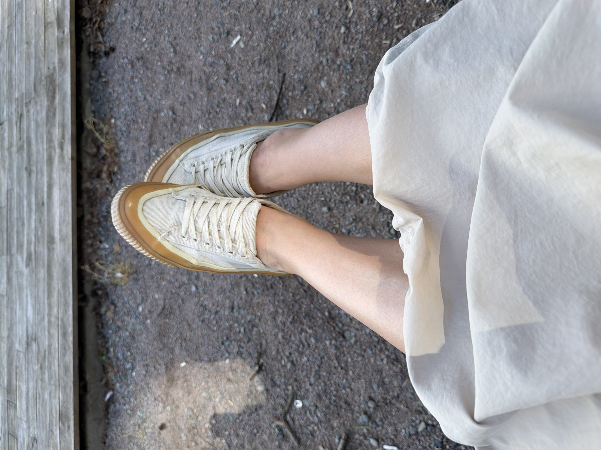 The image shows a top-down view of a person's legs and feet on an outdoor surface. The person is wearing light-colored sneakers with tan rubber soles and laces, and a light-colored skirt or dress. The ground is gravel with some small debris. A wooden structure is partially visible on the left side of the frame. The location is identified as Chaikovsky, Russia.