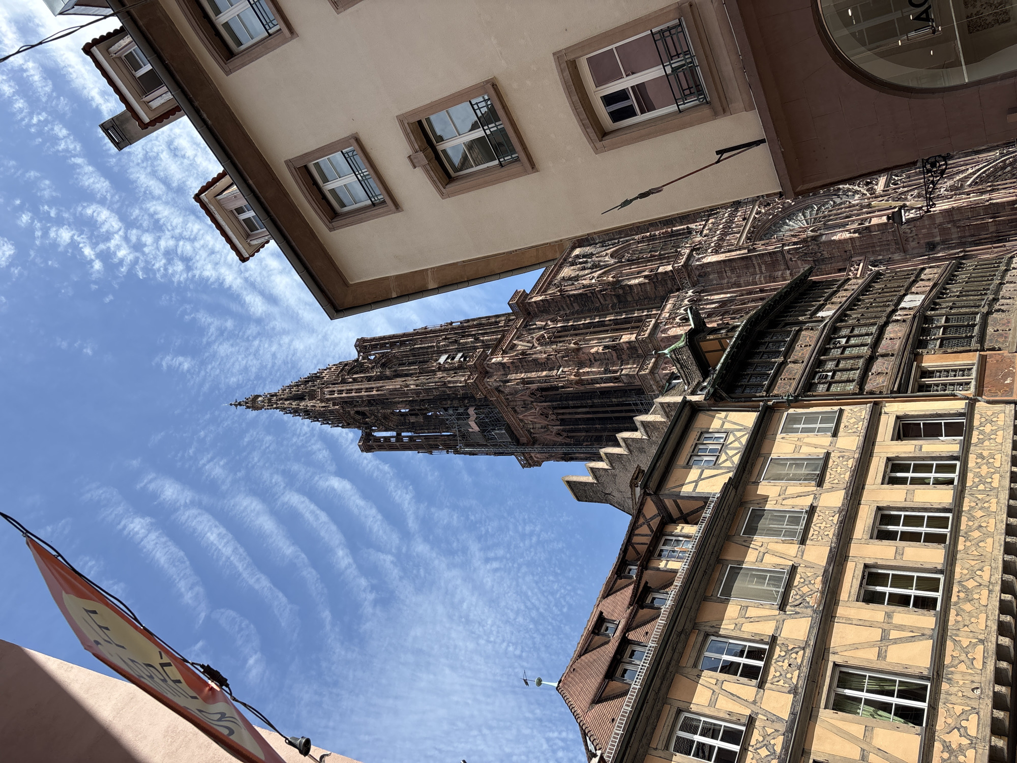 The image shows a low-angle view of a city street in Strasbourg, France, with a prominent Gothic cathedral tower dominating the background. The cathedral, constructed of reddish-brown stone, features intricate architectural details, including spires, buttresses, and arched windows. In the foreground, a row of buildings with traditional Alsatian architecture lines the street. These buildings display half-timbered facades with yellow stucco and dark wooden beams, characteristic of the region. The windows are multi-paned and some have dark metal railings. A bright blue sky with wispy white clouds forms the backdrop. A red and yellow banner with text is visible in the upper left corner of the frame, partially obscuring the sky. The overall scene depicts a sunny day in a historic European city.