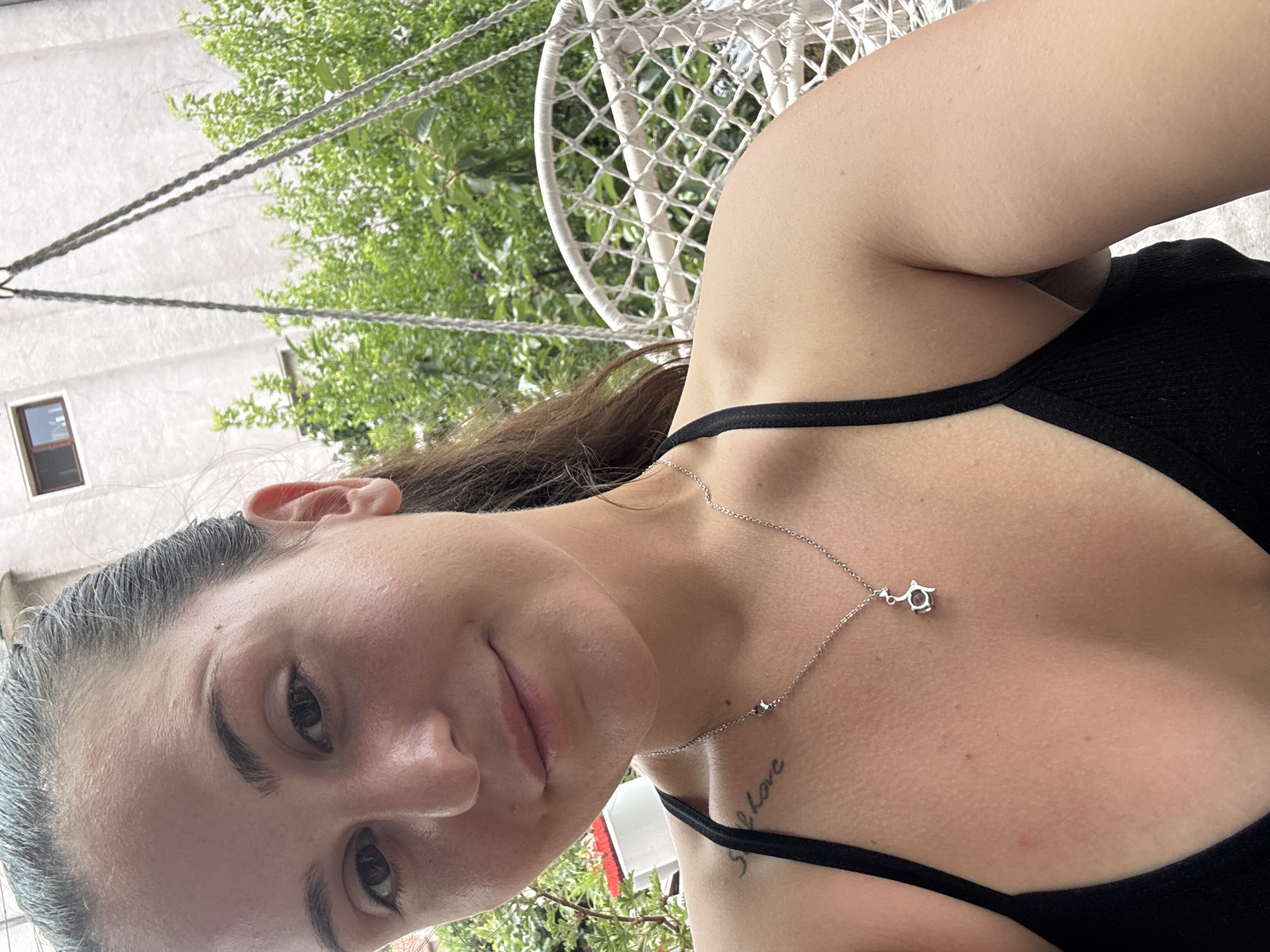 A young woman with dark hair pulled back is captured in a close-up, sideways shot, lying down outdoors. She is wearing a black tank top and a delicate silver necklace with a small pendant. Her gaze is directed towards the camera, and her expression is relaxed, with a hint of a smile.
The setting appears to be a backyard or patio area, with lush green foliage visible in the background. A white, woven hanging chair with a net-like pattern is also present, suggesting a comfortable, leisurely environment. Part of a white building and a window are visible on the left side of the image.
The lighting suggests it is daytime, possibly late afternoon given the soft, diffused light. The weather seems pleasant, conducive to spending time outdoors. There are no discernible clouds in the sky.
A small tattoo is visible on her upper chest, with faint script that reads "I love". The image has a casual, candid feel, as if taken by a friend or herself during a relaxing moment. The overall impression is one of tranquility and ease in an outdoor setting.