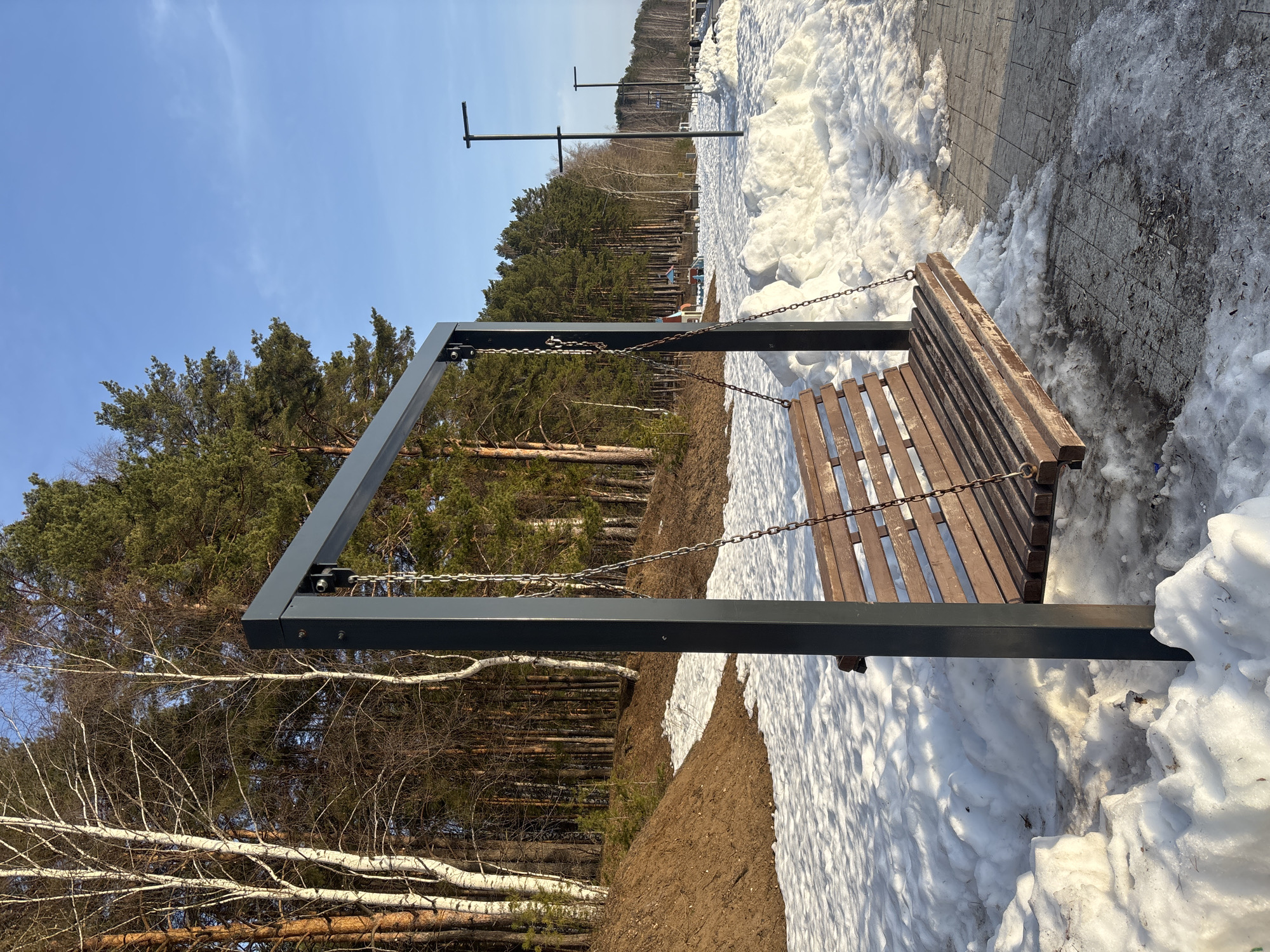 The image shows a wooden swing bench suspended by chains from a gray metal frame, positioned on a snowy landscape in Chaikovsky, Russia. Patches of brown earth are visible where the snow has melted. In the background, there's a dense forest of tall trees, some bare and some with green foliage. Further back, a row of lampposts extends into the distance. The sky above is a clear, bright blue. A paved walkway is visible to the right of the bench.
