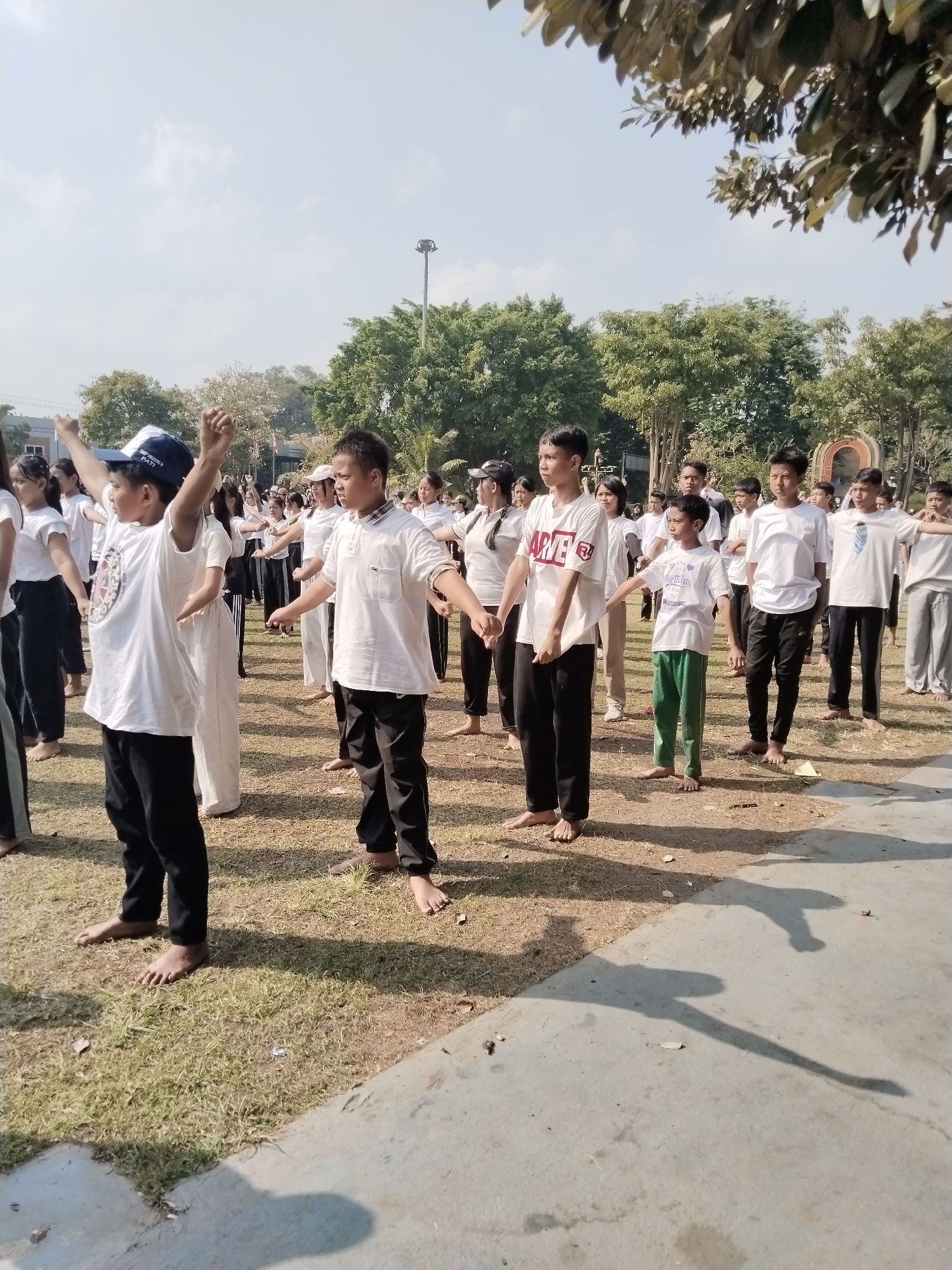 In an outdoor grassy area in Kecamatan Pati, Indonesia, a group of approximately 20 young people, primarily children and adolescents, are engaged in a coordinated physical activity, possibly a form of exercise or martial arts. They are all dressed in casual attire, predominantly white t-shirts and dark pants, with some individuals also wearing white or dark caps. Most of the participants are barefoot. The individuals are arranged in rows, facing forward, with their arms extended and bodies positioned in similar stances, suggesting they are following instructions from an unseen leader. The environment is sunny with a clear blue sky and scattered clouds. Trees and foliage form a backdrop, and in the distance, a lamppost and a decorative archway are visible. The ground is covered with dry grass and some fallen leaves. The scene is captured from a slightly elevated, eye-level perspective, with the shadows of the participants cast on the concrete path in the foreground.