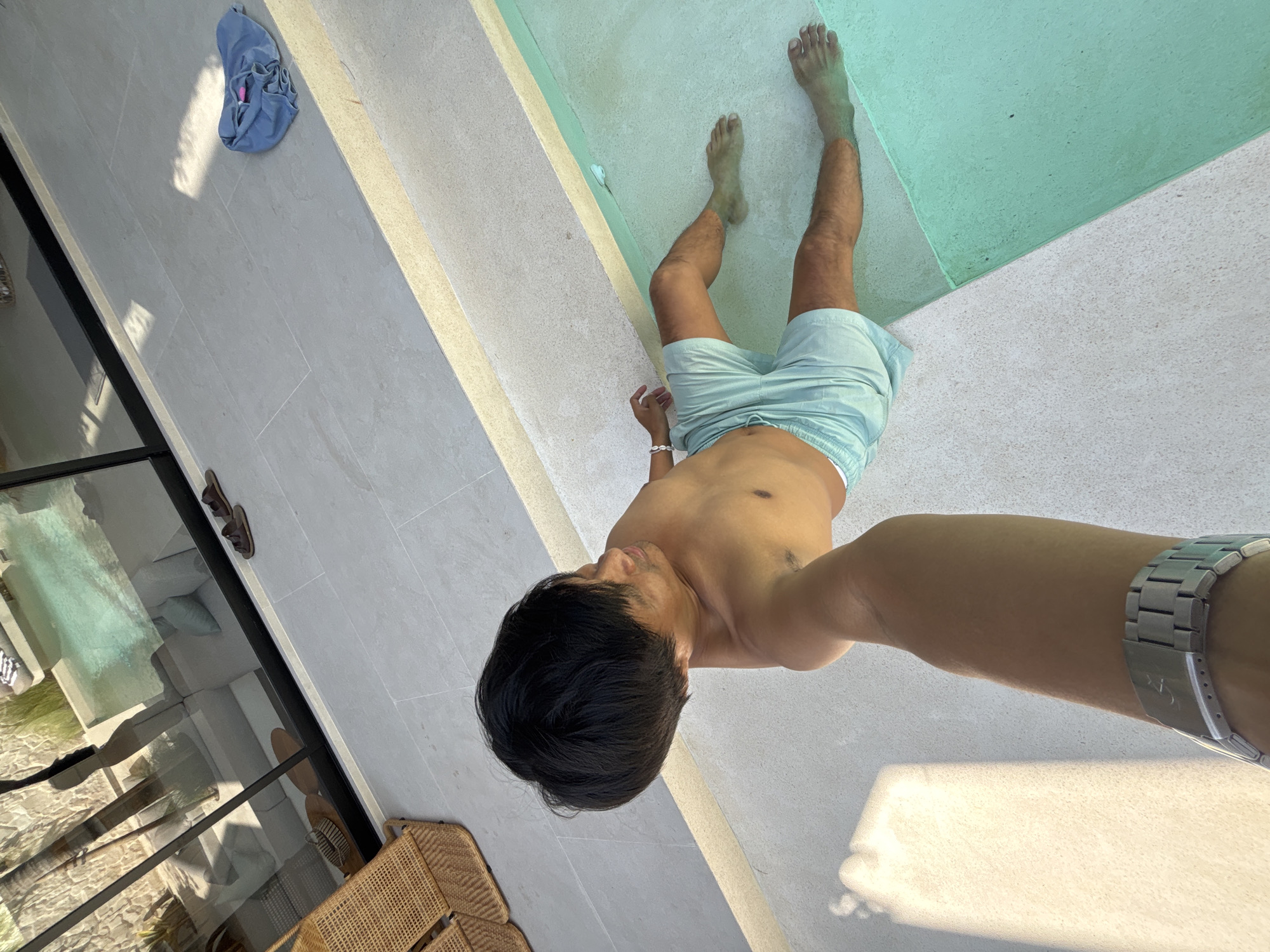 In North Kuta, Indonesia, a person is partially submerged in a turquoise pool, with their legs and lower torso visible in the water. They are wearing light blue swim shorts. The person is positioned on the edge of the pool, with their body angled away from the camera. In the foreground, a forearm with a silver watch on the wrist is visible, suggesting the image is a selfie. The surrounding area appears to be a modern outdoor space with light-colored tiled flooring. Across the pool, through a large glass window, a living area with light-colored furniture and an outdoor chair made of woven material can be seen. A pair of brown sandals and a blue folded garment are present on the tiled surface near the pool.