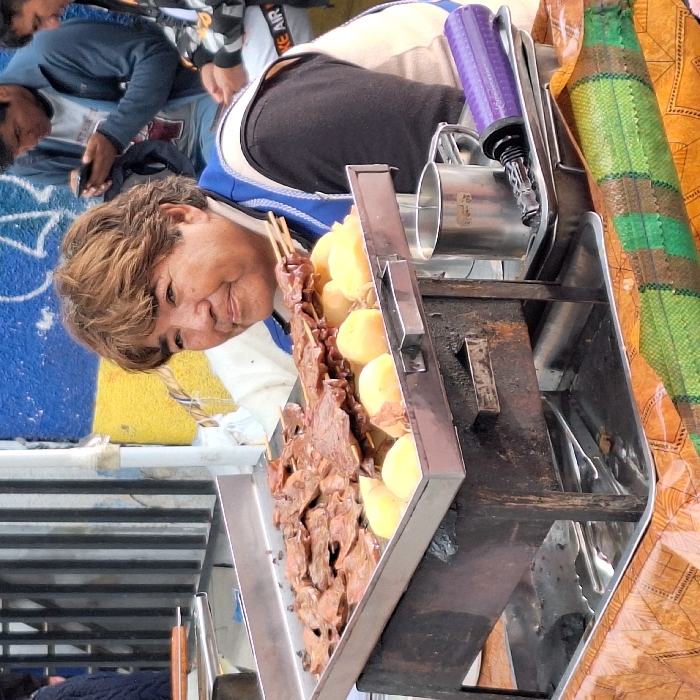 A woman stands beside a food cart displaying skewers of meat and potatoes. The cart features a stainless steel container with grilling implements and a purple pump-like device on top. People are visible in the background, one wearing an “AIR” labeled item. A patterned textile is draped next to the cart.