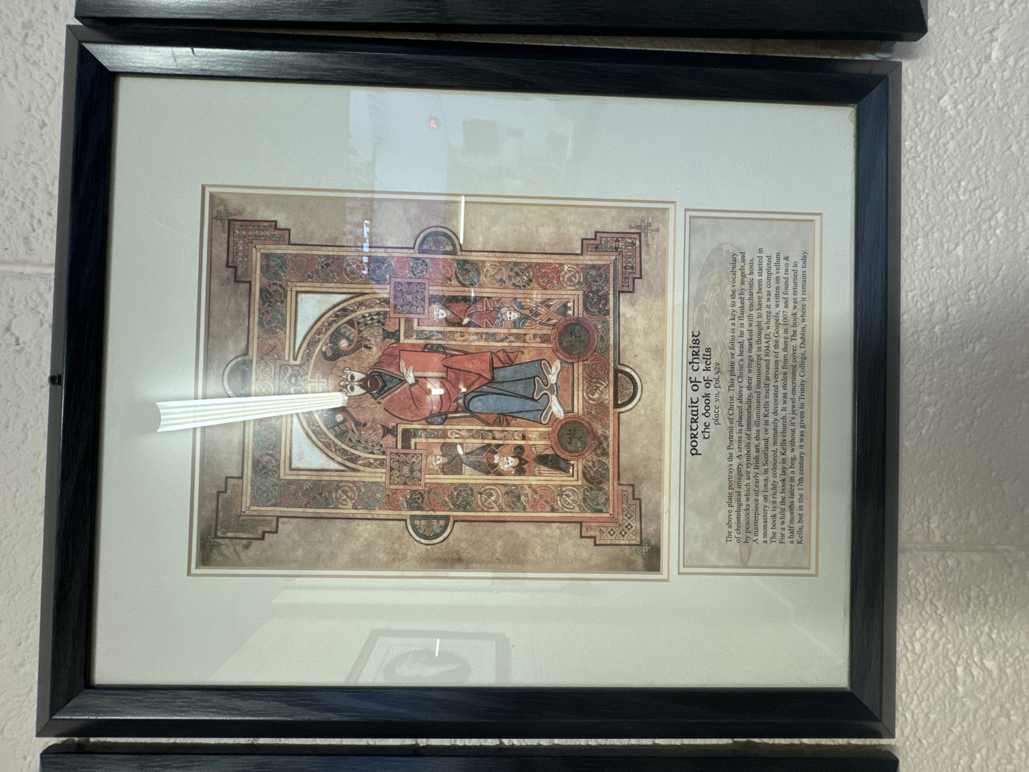 A framed print hangs on a white brick wall. The print depicts an illuminated manuscript page featuring a central figure of Christ with a beard and crown, dressed in red robes and blue trousers. The figure is seated and flanked by smaller figures and decorative borders with intricate knotwork and animal motifs. Below the illustration is a descriptive text box. The scene is lit by overhead lighting, which causes a glare on the glass of the frame. The frame is black and has a textured finish. Two other framed items are partially visible to the left and right of the central frame, also appearing to be prints. The overall setting suggests an interior space, possibly a museum, gallery, or educational institution. There is no indication of the city of Waterford, Ireland, or any specific location within it.