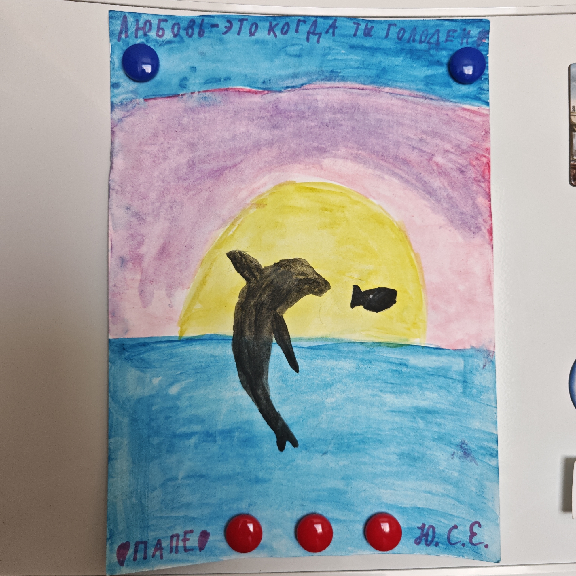 A child's drawing is affixed to a white refrigerator in Taganrog City District, Russia. The artwork, executed in watercolor, depicts a dolphin leaping toward a fish against a backdrop of a yellow sun setting into a blue sea. The sky is rendered in shades of pink and purple. The text "Любовь - это когда ты голоден" (Love is when you are hungry) is written across the top. The drawing is attached to the refrigerator with two blue magnets at the top and three red magnets at the bottom. The name "ПАЛЕО" and "Ю.С.Е." are written in the bottom corners of the drawing.