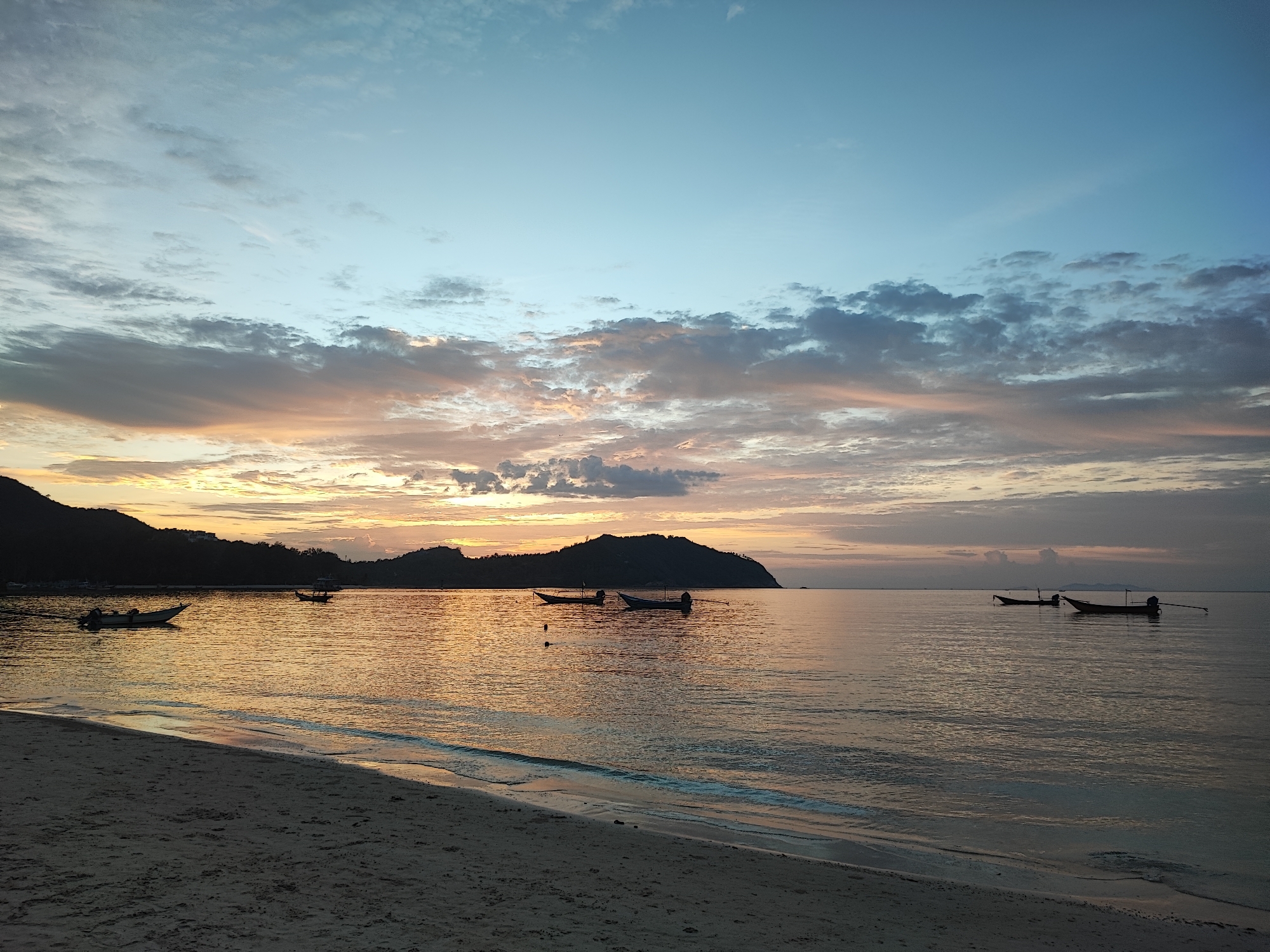 The image depicts a serene beach scene at dusk in Ko Pha-ngan District, Thailand. The sky transitions from a clear, pale blue at the top to hues of orange, pink, and gray clouds near the horizon, indicating either sunrise or sunset. The calm sea reflects the colors of the sky, creating a shimmering effect. Several longtail boats are visible on the water, some moored and others seemingly abandoned. In the background, silhouetted hills and a shoreline are dimly visible. The foreground features a sandy beach with gentle waves lapping at the shore. There are no people visible in the image. The overall atmosphere is tranquil and peaceful.