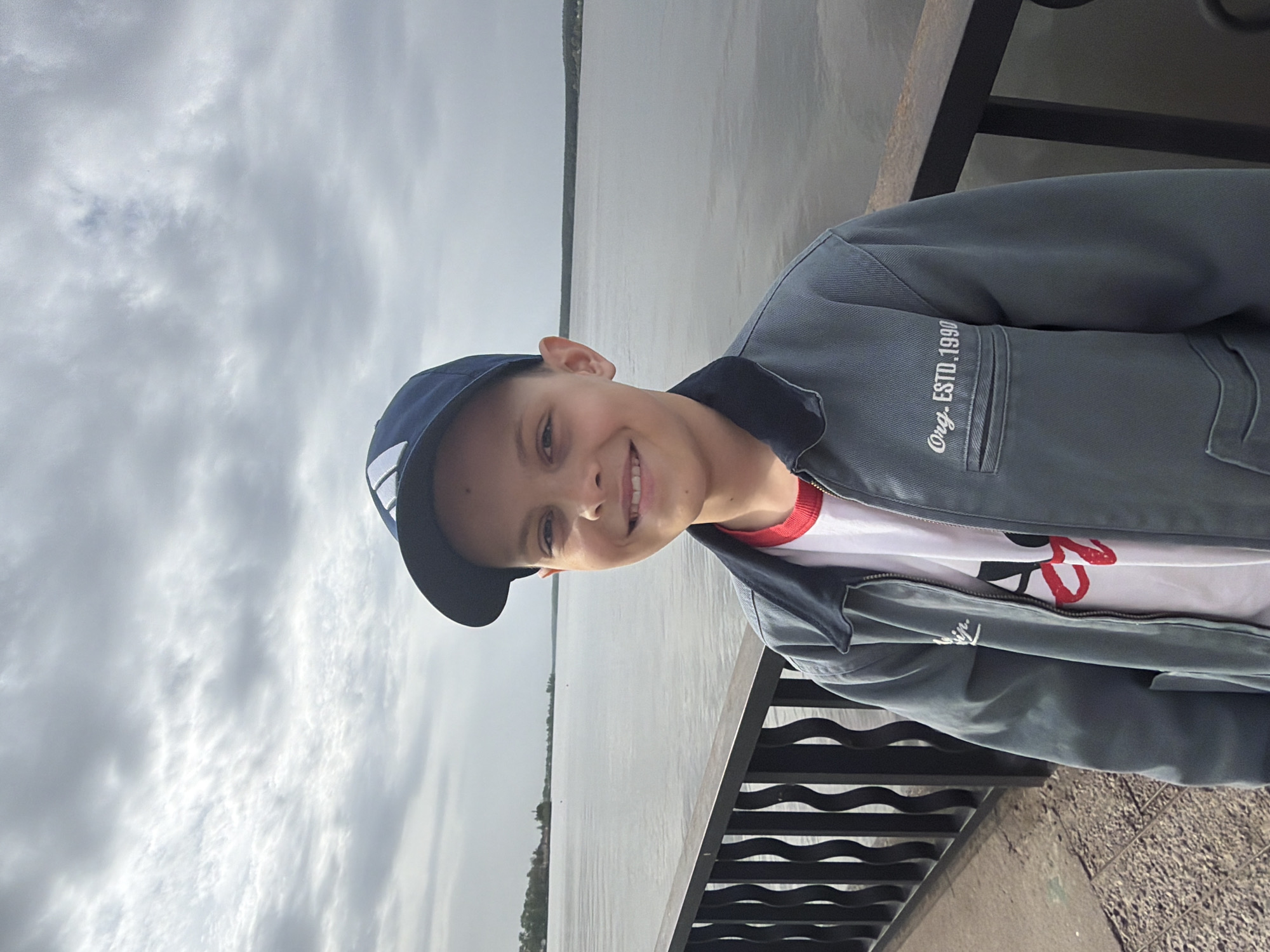 A young boy wearing a blue baseball cap and a gray jacket with the words "Org. ESTD. 1990" on the chest pocket is standing on a bridge or elevated walkway with a black metal railing. In the background is a wide body of water, likely a river or reservoir, with a strip of land visible on the opposite shore. The sky is overcast with gray clouds. The image suggests an outdoor setting, potentially near a body of water in a city. It is impossible to confirm that this is a location in Votkinsk, Russia without more information.