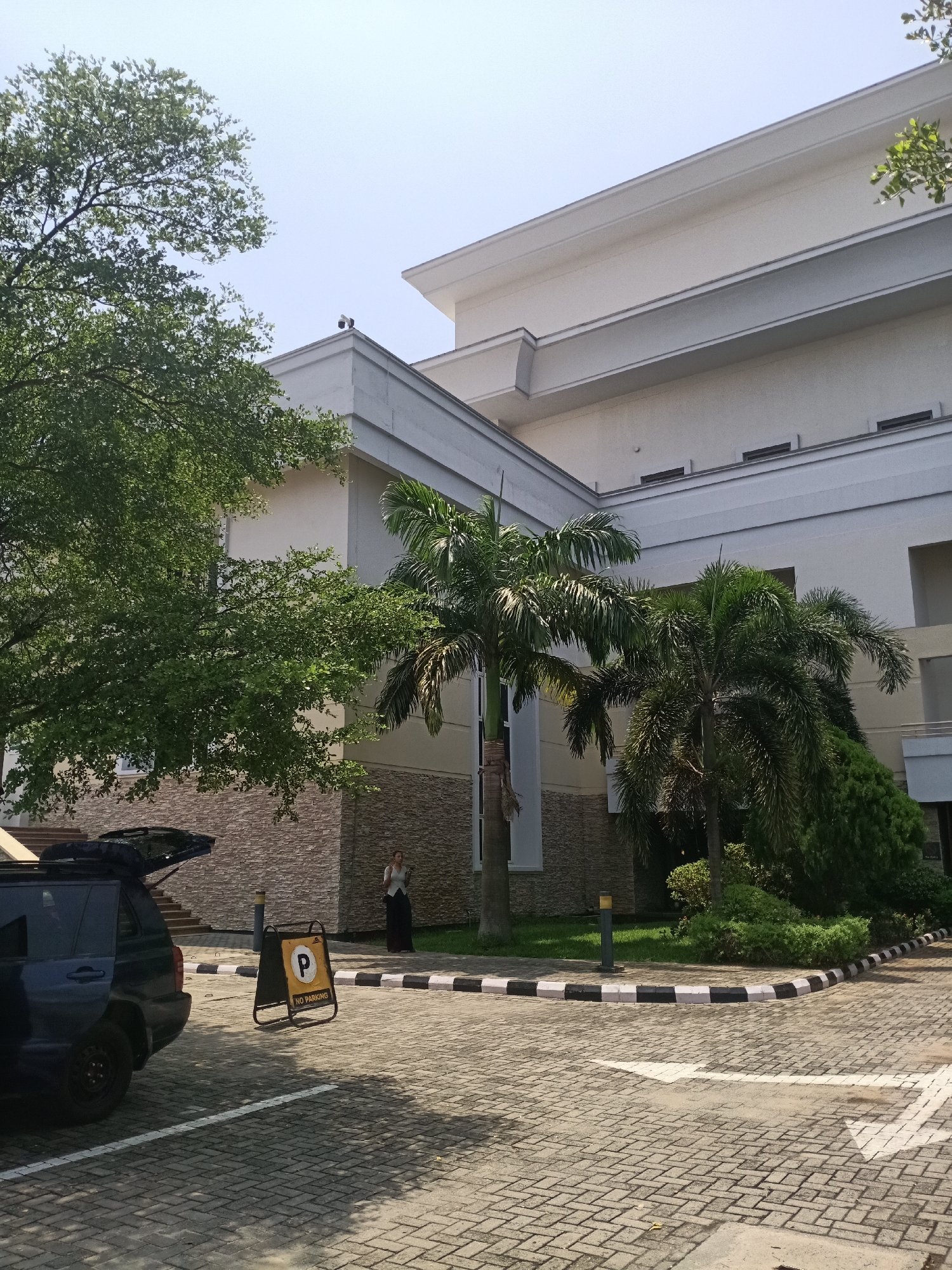 A sunny, daytime shot shows a multi-story, light-colored building with palm trees and landscaping in Igboefon, Nigeria. The building has a combination of stone and white siding. A black SUV with the trunk open is parked in the foreground near a "No Parking" sign. A woman is standing near the building. A white arrow is painted on the brick-paved driveway, indicating the flow of traffic. A security camera is visible on the roof of the building.