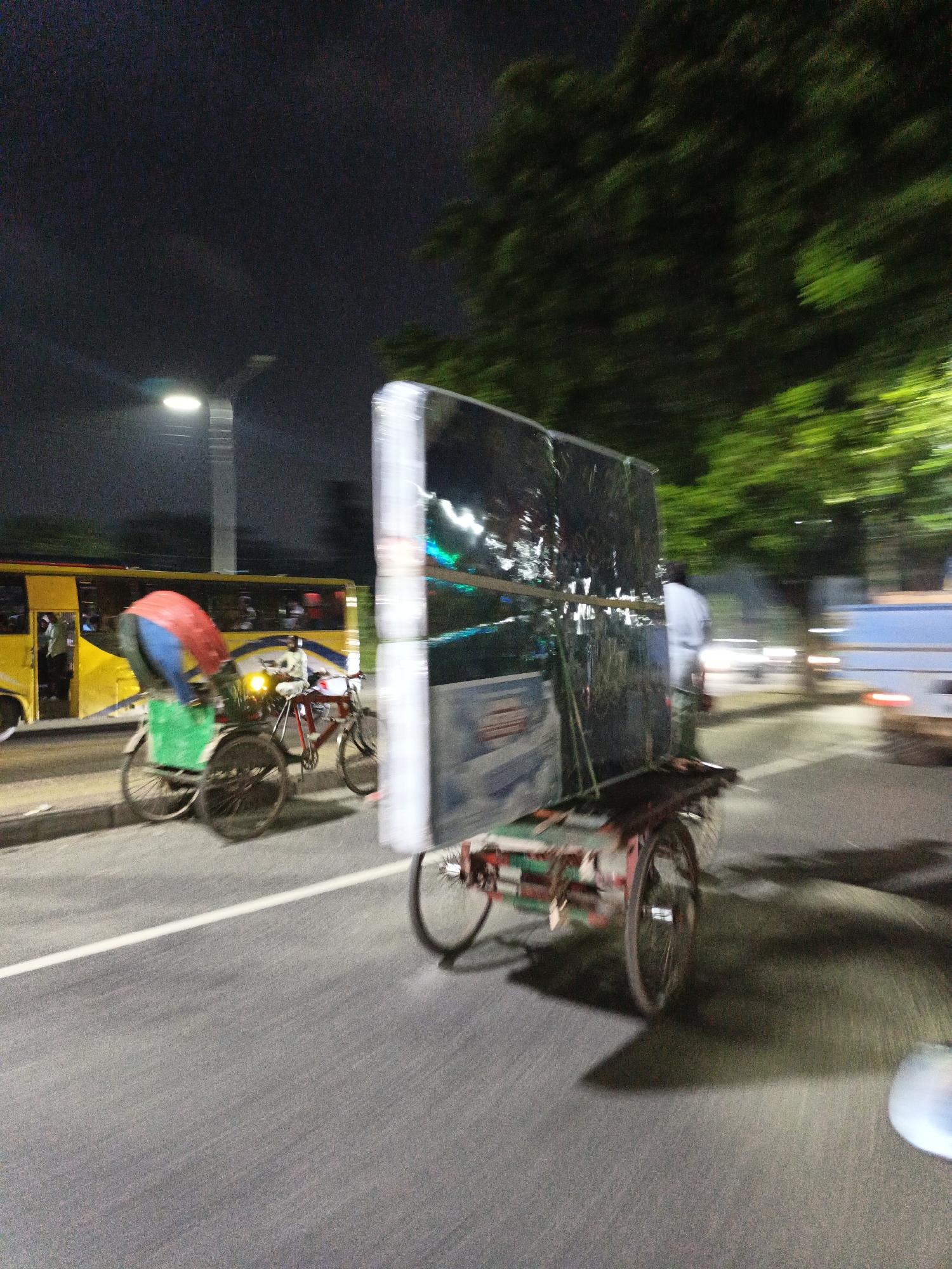 The image captures a dynamic nighttime scene in Dhaka, Bangladesh, likely on a busy street. In the foreground, a cycle rickshaw is in motion, carrying a large, rectangular object wrapped in plastic sheeting. The object appears to be a large flat-screen television or similar electronic appliance, secured with ropes and strapped to the rickshaw's platform. The rickshaw has two large wheels at the back and one smaller wheel at the front, typical of this mode of transport. The driver, wearing a light-colored shirt, is partially visible, pedaling to move the rickshaw forward.
To the left of the rickshaw, a yellow and blue bus is also in motion, its headlights and interior lights illuminating the scene. Passengers are visible inside the bus. A street light with a circular lamp is visible in the background, casting a bright light on the road and surrounding area. Trees with blurred green foliage are visible on the right side of the image, suggesting a roadside environment. The overall impression is one of a bustling urban environment at night, with vehicles and people moving along a paved road. The motion blur indicates a relatively slow shutter speed, capturing the movement of the subjects.
