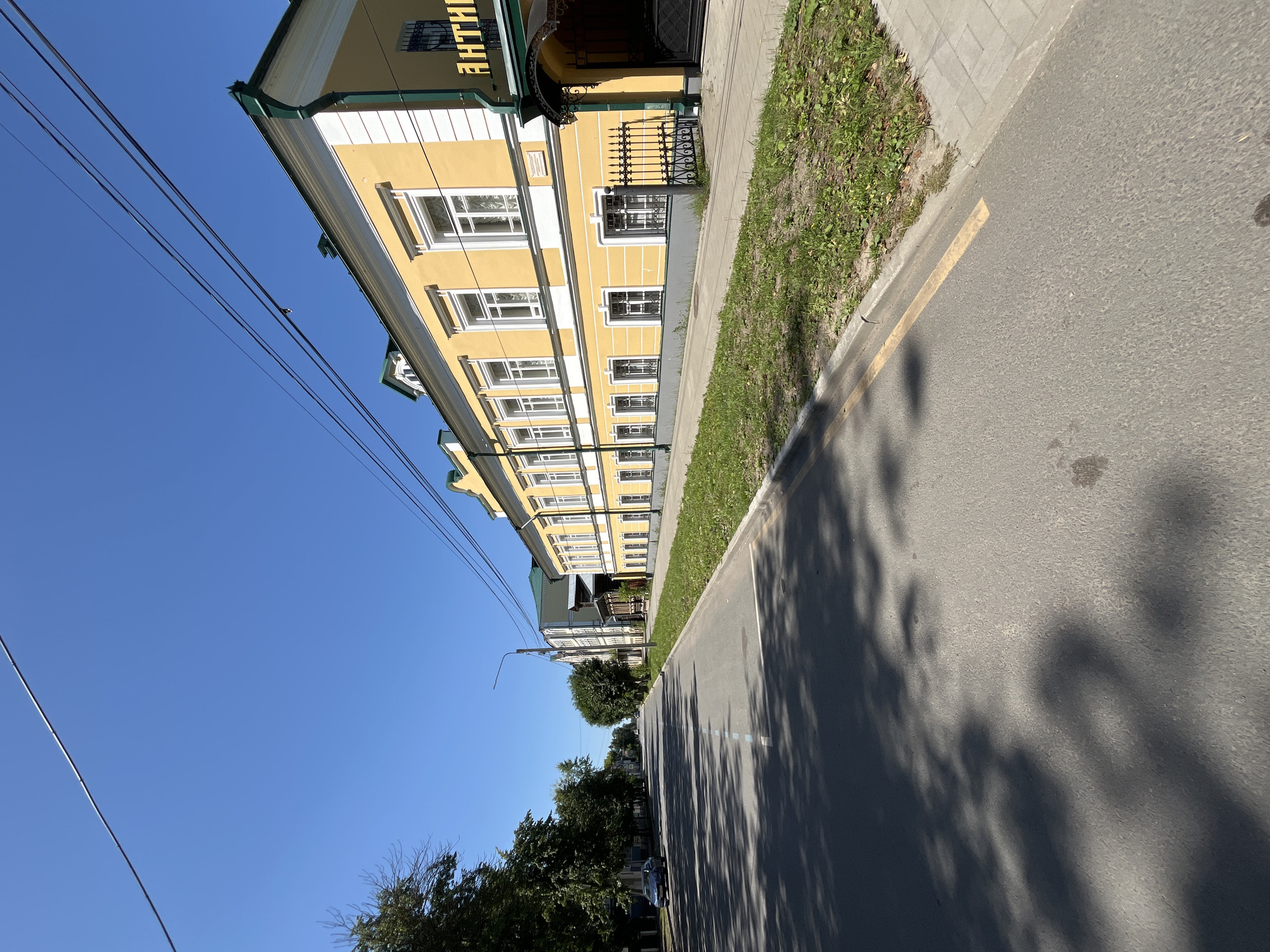 A street scene in Kladovitsy, Russia, on a clear, sunny day. The perspective is from the roadway, looking down a paved street lined with buildings and trees. On the right side of the street, a long, two-story yellow building with white trim and green accents is prominent. It has multiple windows on both floors, many with decorative white shutters. An awning with the Cyrillic word "АНТИК" (likely meaning "antique") is visible over an entrance. A black metal fence separates the building from the sidewalk. On the left side of the street, a line of mature trees casts shadows across the asphalt. A blue car is parked on the left side of the road. The sky is a bright, cloudless blue. Overhead power lines are strung across the scene. The roadway has painted lane markings and a yellow line along the curb. The sidewalk on the right is paved with rectangular stones. The environment appears to be a quiet, residential or commercial street within a town or city.