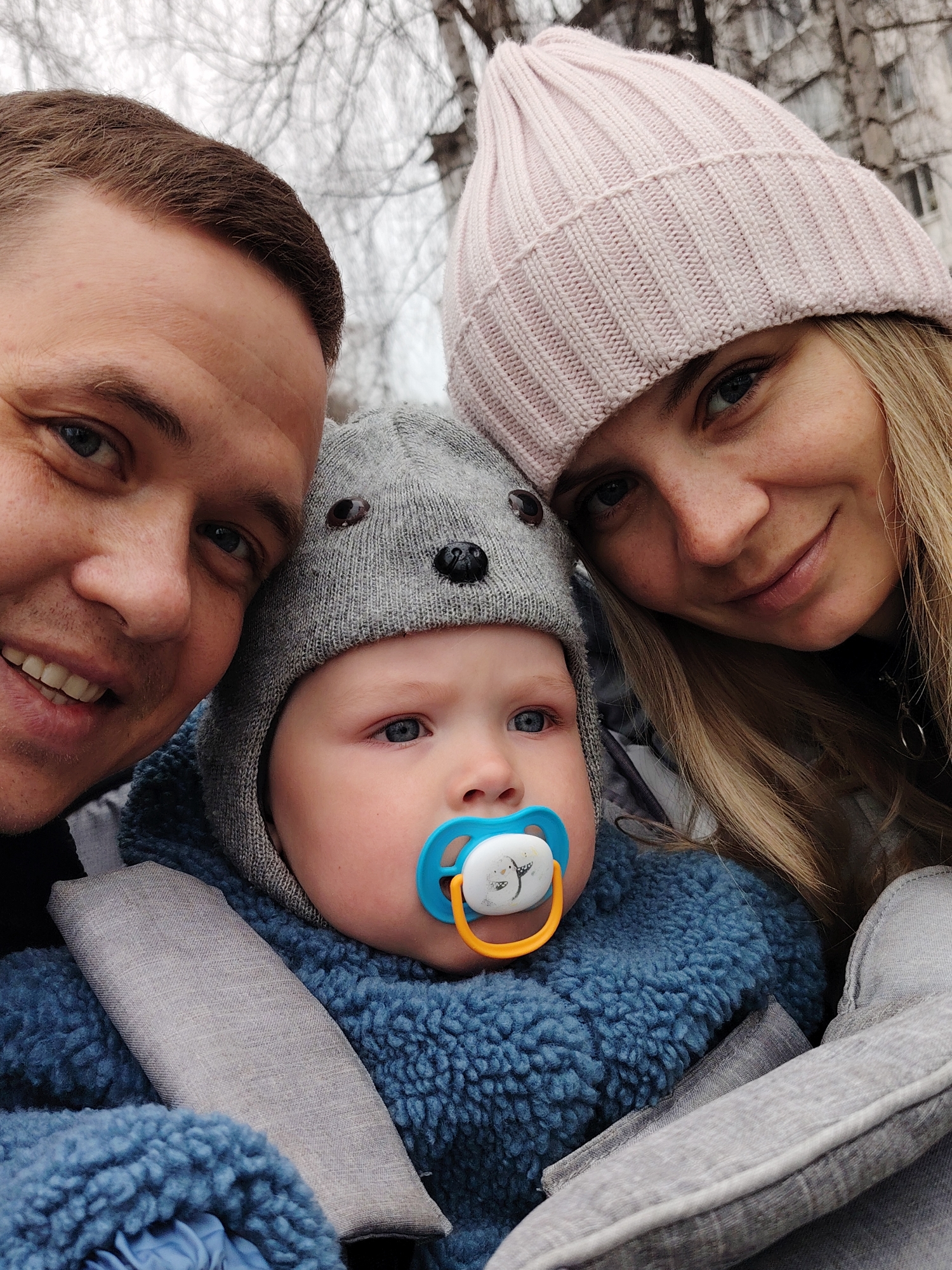 A close-up, overhead selfie captures a family of three outdoors, likely in Chaikovsky, Russia, during cool weather. The image prominently features a man on the left, a woman on the right, and a baby nestled between them in the center.
The adult male, positioned closest to the camera on the left, has short brown hair and blue eyes, and smiles broadly. The adult female on the right also smiles, with long blonde hair visible beneath a light pink ribbed knitted beanie. Both adults appear happy and engaged, their faces filling the top and sides of the frame.
The baby, situated centrally, wears a charming grey knitted hat with small bear ears and a blue, textured, sherpa-like outerwear. A blue and yellow pacifier, adorned with a small penguin-like illustration on its white disk, is in the baby's mouth. The baby's bright blue eyes are open, looking slightly upwards with a curious or calm expression. The baby appears to be secured in a stroller or car seat, indicated by grey fabric straps and padding visible around its chest and side, suggesting an outing.
The background shows bare tree branches against an overcast sky, suggesting a cold season like winter or early spring. A hint of a building can be seen behind the woman's head in the distance. The overall mood is warm and familial, despite the cold outdoor setting. No discernible text is visible in the image.