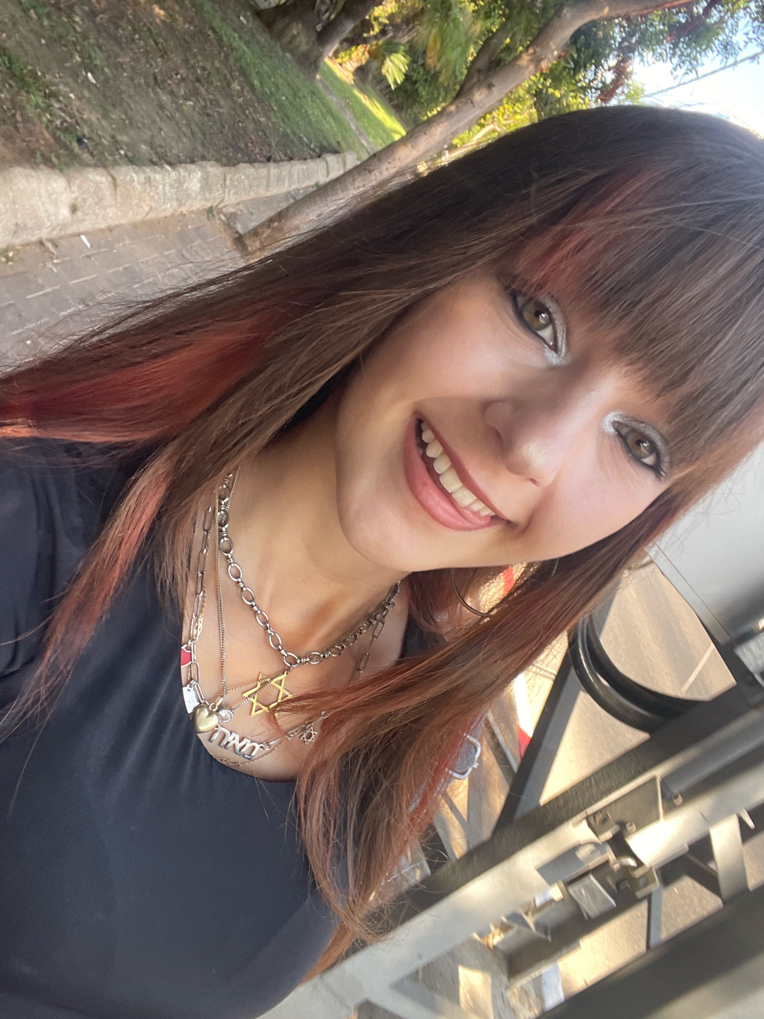 A young woman with brown hair, streaks of red, and bangs is taking a selfie outdoors. She is wearing a black top and several necklaces, including one with a Star of David pendant and another with Hebrew lettering. Her makeup includes silver eyeshadow and dark eyeliner. She is smiling, with her tongue slightly sticking out. The background shows a pathway, grass, trees, and sunlight, suggesting a park or street in Raanana, Israel.