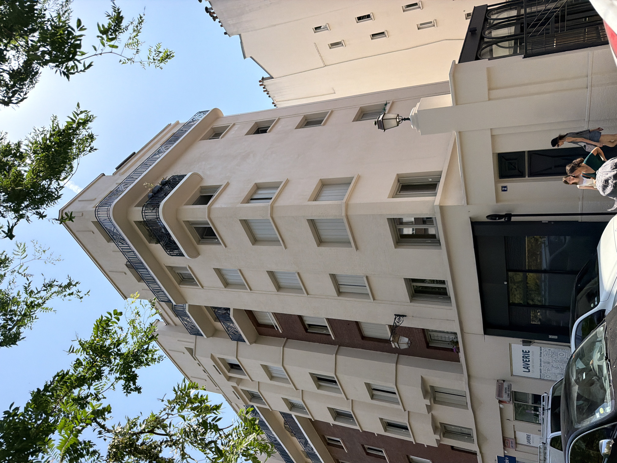 This is a low-angle, slightly tilted outdoor photograph of a multi-story building under a clear blue sky. The building appears to be residential or commercial, with a light-colored facade featuring numerous windows and balconies. Some of the windows are small and rectangular, while others are larger and set within recessed frames. The balconies have decorative black metal railings. A portion of the building's facade is clad in dark red brick.
In the foreground, the branches of green leafy trees obscure parts of the sky and the building. The scene suggests it is daytime, with bright sunlight illuminating the building. A few people are visible near the ground level of the building, walking or standing. One person is wearing a dark vest over a lighter top and a skirt, another has on a green top and shorts, and a third is in a white top and skirt.
On the ground floor, there are signs indicating a "LAVERIE" (laundry) with additional text in French: "LIBRE-SERVICE" (self-service), "coparte-yeties court-Séchage rapide" (washing machines - short - quick drying). There are also cars parked in front of the building, one of which is partially visible on the right side of the frame. The overall impression is a street scene in an urban environment in France.