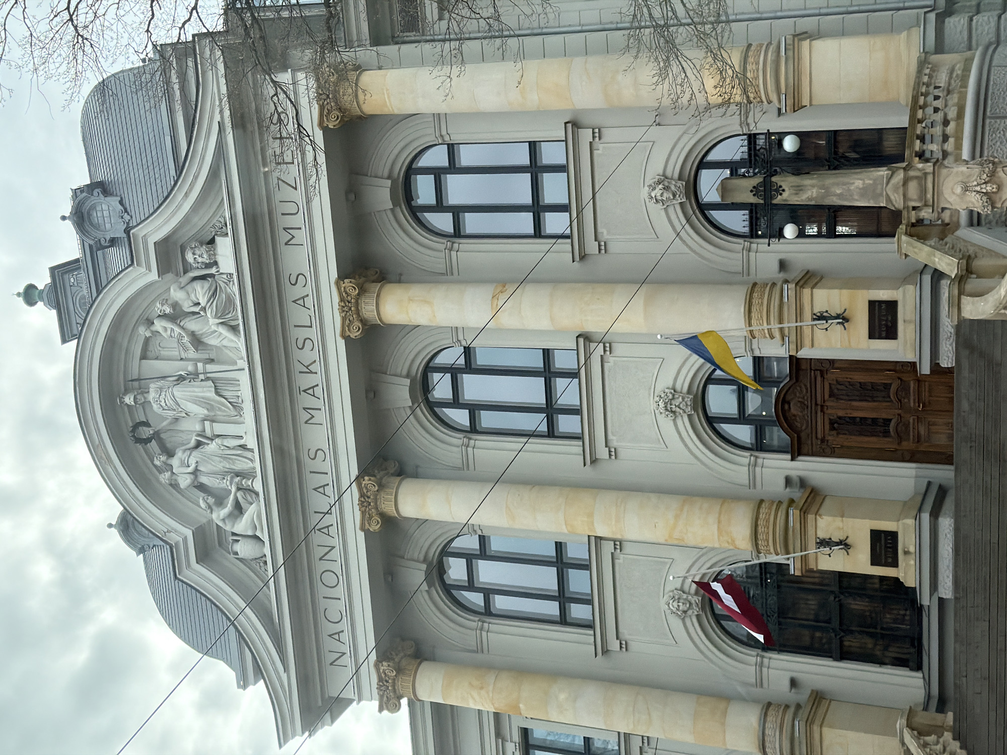 The image presents a grand, neoclassical building facade, identified as the Latvian National Museum of Art (Nacionalais Makslas Muzejs) in Riga, Latvia, under an overcast sky. The stately structure features a light-colored stone exterior with prominent, cream-colored fluted columns topped with ornate Corinthian capitals, spanning multiple floors.
Above the main entrance area, an elaborate frieze showcases classical-style sculptures of human figures, some draped and others partially nude, seemingly depicting artistic or allegorical themes. The museum's name, "NACIONALAIS MAKSLA MUZEJS," is carved horizontally into the building's facade, positioned beneath the frieze and above a series of arched windows with dark frames. Smaller decorative carved heads are also visible between some windows.
At street level, near a set of large, dark wooden double doors, two flags are displayed. The upper flag is the blue and yellow of Ukraine, while the lower flag is the maroon and white of Latvia. A dark plaque with the word "MUSEUM" is visible near one of the flagpoles. Bare tree branches extend into the frame against the cloudy sky, suggesting a cold season such as late winter or early spring. The scene is static, depicting the building's exterior in diffused daylight.