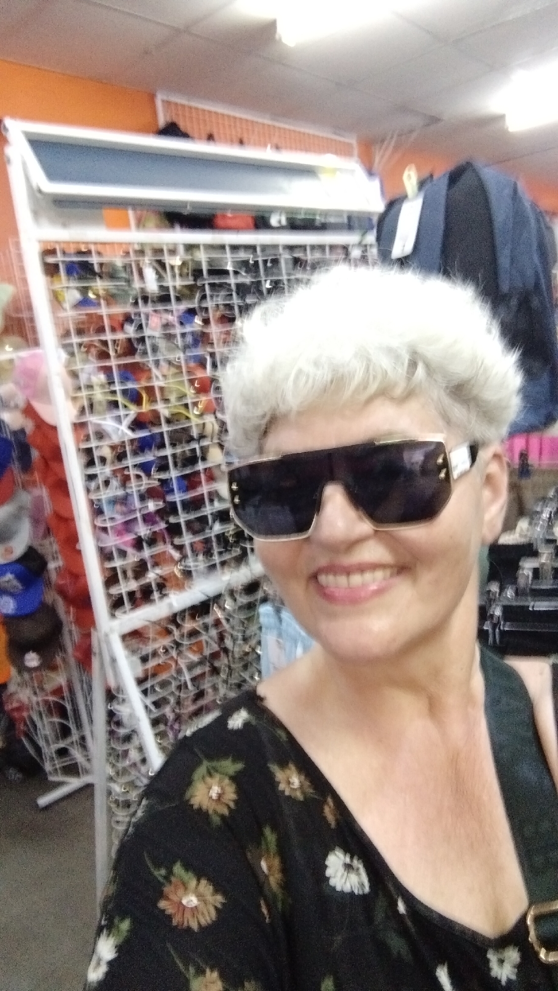 In the city of Horishni Plavni, Ukraine, a woman with short, white, curly hair is pictured indoors. She is wearing large, dark-lensed sunglasses with gold accents and a black top with a floral pattern. She is smiling. Behind her, a rack filled with sunglasses is visible, and to her left, a display of hats can be seen. A dark backpack is also visible in the background. The environment appears to be a retail store.
