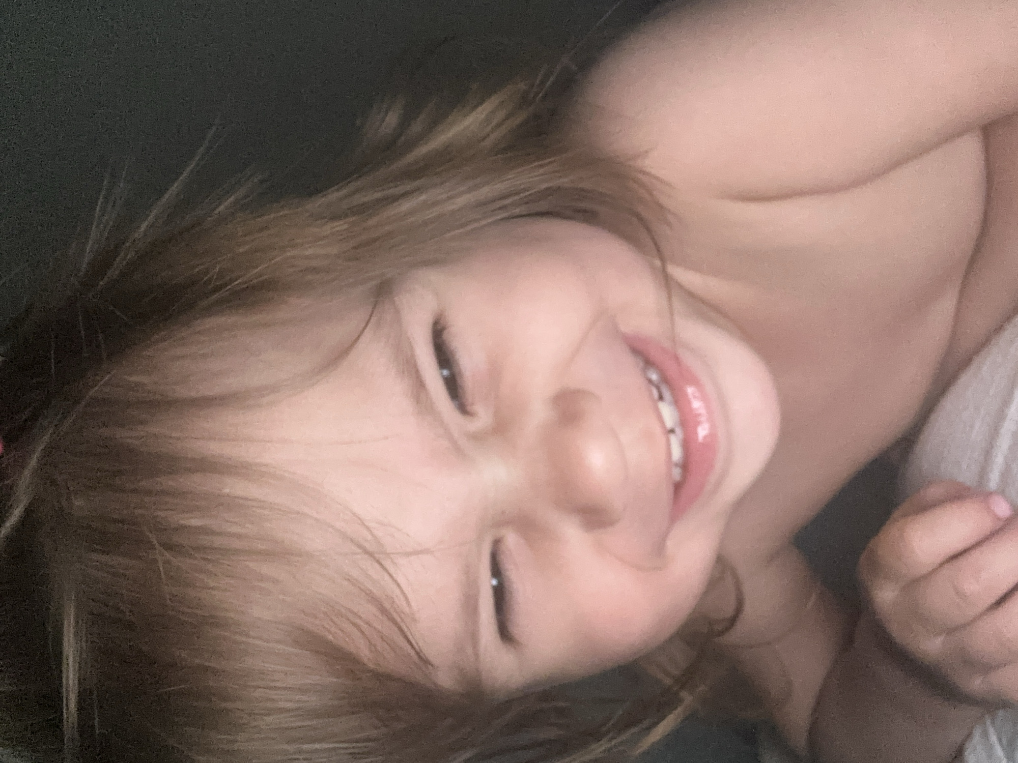 The media file shows a close-up, upside-down, and slightly angled shot of a young child's face. The child has light brown, tousled hair and is smiling broadly, showing teeth that appear to have braces. Their eyes are partially closed, and their cheeks are rosy. The lighting is soft, suggesting an indoor setting, possibly at dawn or dusk, or in a dimly lit room. The background is dark and indistinct, with a hint of white fabric visible on the right, possibly a blanket or clothing. The child's bare shoulder and arm are also visible, indicating they are likely undressed or wearing minimal clothing. The overall impression is one of happiness and innocence. There is no visible text in the image.