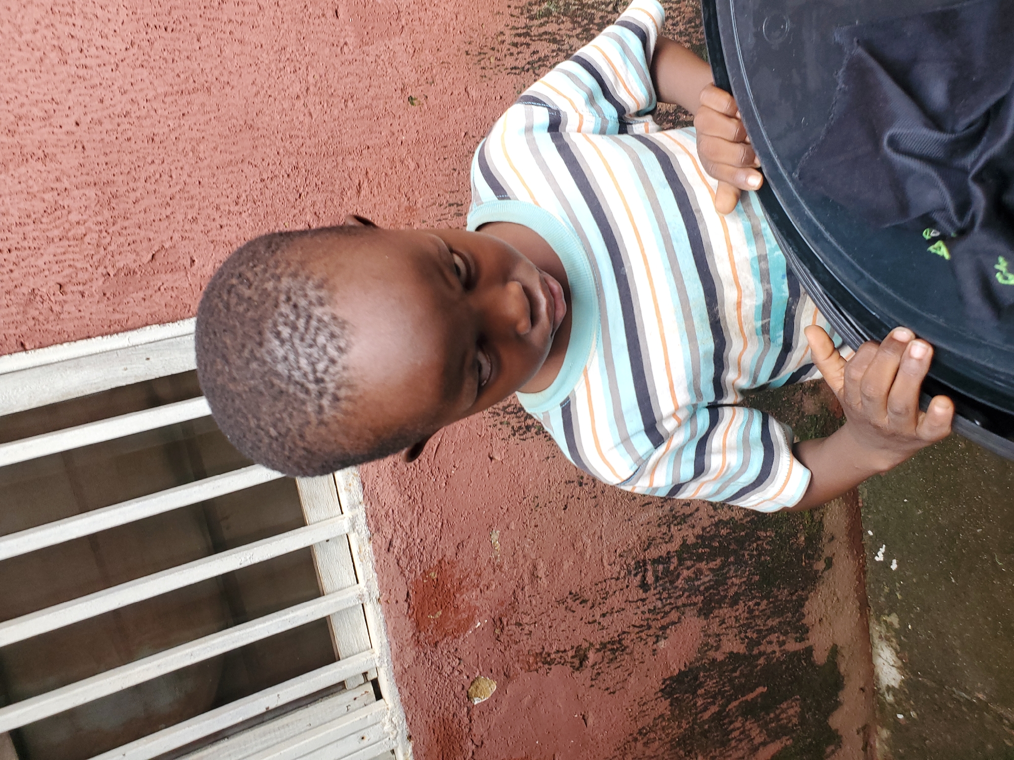 A young child, appearing to be a boy, is the sole subject in this image. He is wearing a striped t-shirt with vertical bands of light blue, white, grey, and orange. The child is positioned in what appears to be an outdoor setting. To his left, there is a light-colored metal grate or vent system set into a rough, reddish-brown textured wall. To his right, a large, black object, possibly a barrel or container with a lid, is visible. The child's hands are gripping the edge of this black object. The ground appears to be wet and stained in places with dark, mossy patches. The overall impression is of a somewhat weathered or utilitarian environment. The lighting suggests it could be daytime, but the specific time is not discernible. No text is visible in the image. The child's facial expression is somewhat serious or contemplative, with his gaze directed downwards. The image is oriented such that the child and the surrounding elements are tilted.