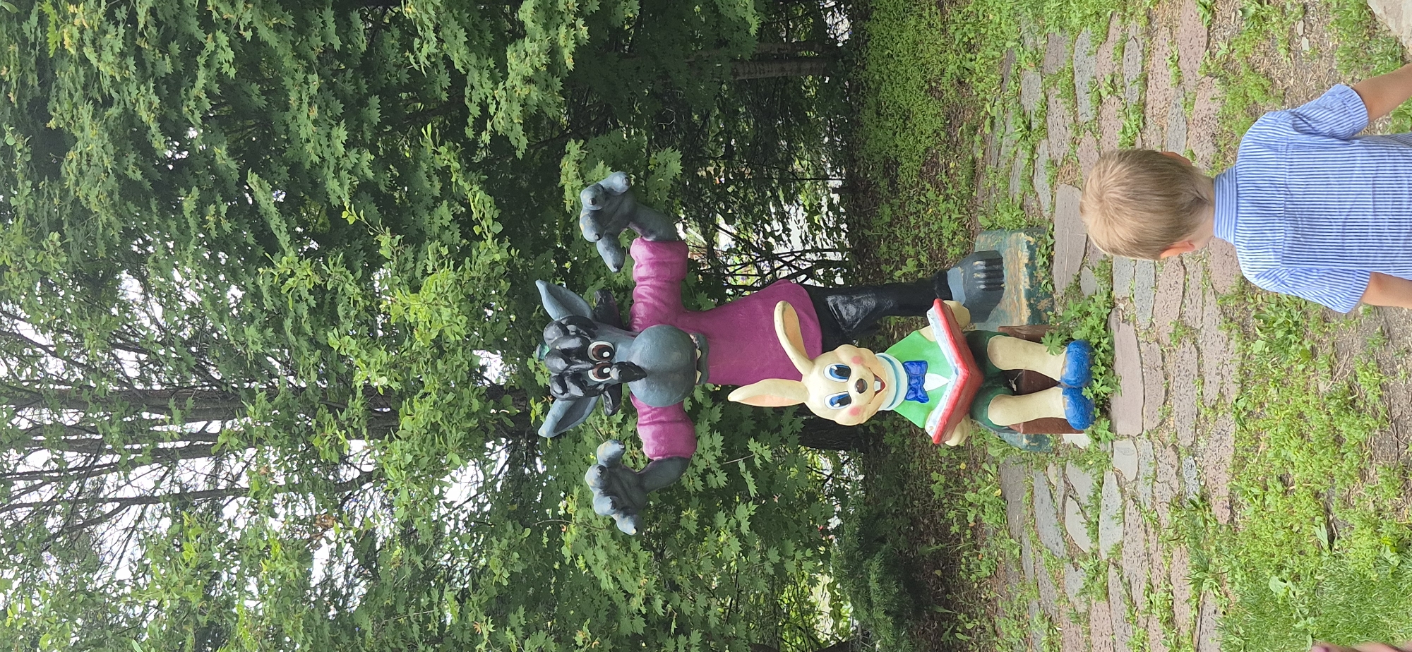 A young boy, seen from behind and at an angle, is standing on a pathway made of irregular stones. He is wearing a blue and white striped shirt. To his left, there are two large, colorful statues. The statue on the left depicts a grey wolf-like character with a prominent snout, large ears, and wide eyes, dressed in a pink shirt and black pants. Its arms are raised, with its hands open. The statue on the right is of a light-colored rabbit character with long ears, wearing a green shirt and blue bowtie. The rabbit is holding an open red book. The statues are positioned on a base, with the rabbit seemingly standing on the wolf's outstretched foot. The scene is set outdoors, with lush green trees and foliage surrounding the pathway and the statues. The lighting suggests it is daytime, with no specific time of day being determinable. The environment appears to be a park or garden area. The boy's presence suggests he might be observing or interacting with the statues. The overall mood is peaceful and perhaps whimsical due to the cartoonish nature of the statues. There is no visible text in the image.