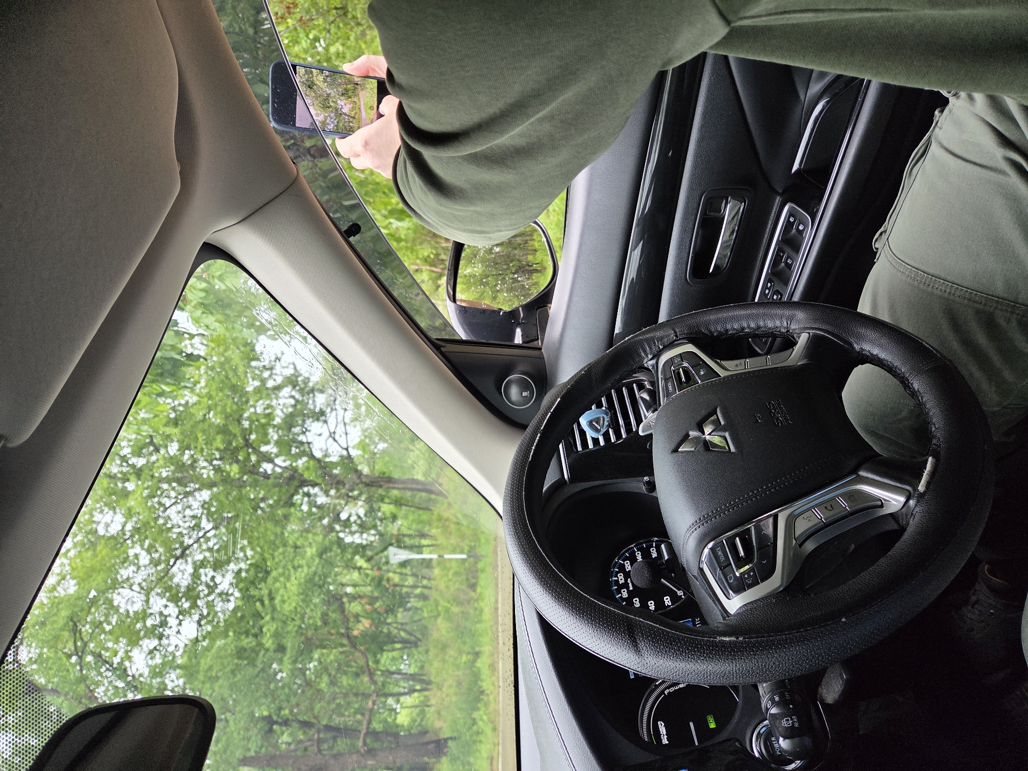 The image displays the interior of a stationary Mitsubishi Outlander PHEV, situated in Pronkino, Russia. The perspective is from the driver's side, looking forward and towards the passenger compartment. A single individual, presumed to be the driver, is visible, wearing an olive-green long-sleeved top and matching cargo-style pants. Their right arm is extended through the open passenger window, holding a dark smartphone horizontally with both hands, appearing to photograph the exterior. Water droplets are visible on the phone's screen.
The vehicle's dashboard indicates a speed of 0 km/h, a "READY" status light for the hybrid system, and an external temperature of 11°C. A blue clip, possibly an air freshener, is attached to a central air vent. The steering wheel, featuring a Mitsubishi logo and integrated controls, is prominent in the foreground. Water droplets are present on the exterior of the windshield and the passenger side window, suggesting recent or ongoing rain.
Beyond the vehicle, a dense, green forest with numerous deciduous trees is visible under an overcast sky. A road or path runs through this natural environment, and a white, inverted-Y shaped road sign stands on the right side of the visible outdoor scene. The passenger side mirror reflects a similar treed landscape.