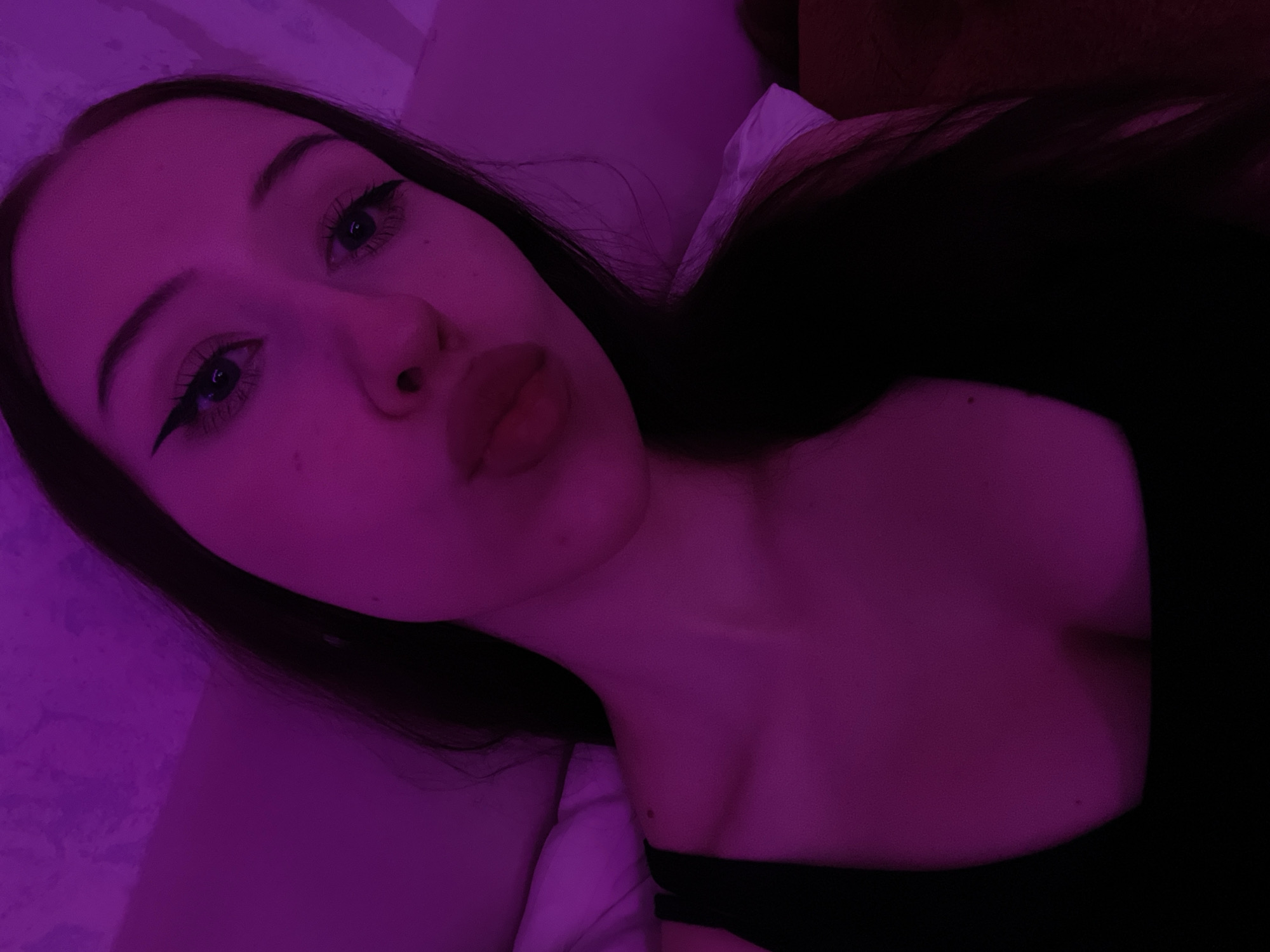 A young woman with dark hair and full lips is seen in a close-up, selfie-style shot. She is wearing a black top and her face is angled towards the camera, with her eyes looking directly into the lens. Her makeup includes winged eyeliner and defined eyebrows. The lighting in the image is predominantly purple, casting a warm glow over her skin and the background, which appears to be a wall or textured surface. The overall setting suggests an indoor environment, possibly a bedroom or a dimly lit room. The photograph does not contain any specific identifiable landmarks or elements that would allow for the determination of its location within the city of Votkinsk, Russia.