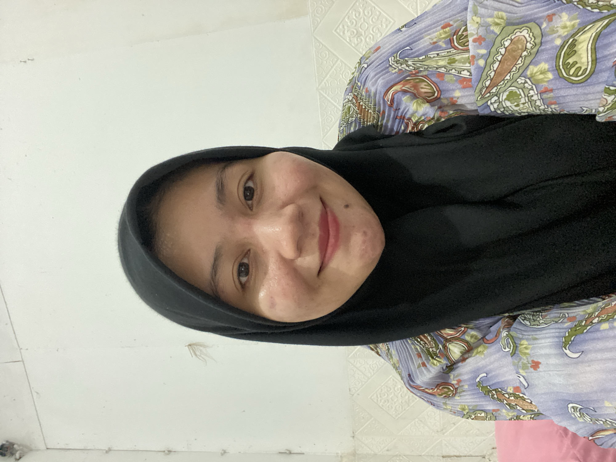 This is a portrait-oriented, close-up, slightly tilted, indoor selfie of a young woman wearing a black hijab and a patterned blouse. The woman is the central subject, with her face filling a significant portion of the frame. She is looking directly at the camera with a slight smile. Her skin has some blemishes and freckles visible. The background is a plain white wall, likely in a home setting, with a textured wallpaper pattern visible on the right side of the frame. The lighting appears to be artificial and somewhat diffuse, suggesting an indoor environment. The overall mood is casual and relaxed. There is no visible text in the image. The clothing of the woman suggests a modest style, and the patterned blouse has floral and paisley motifs in shades of purple, green, and orange.