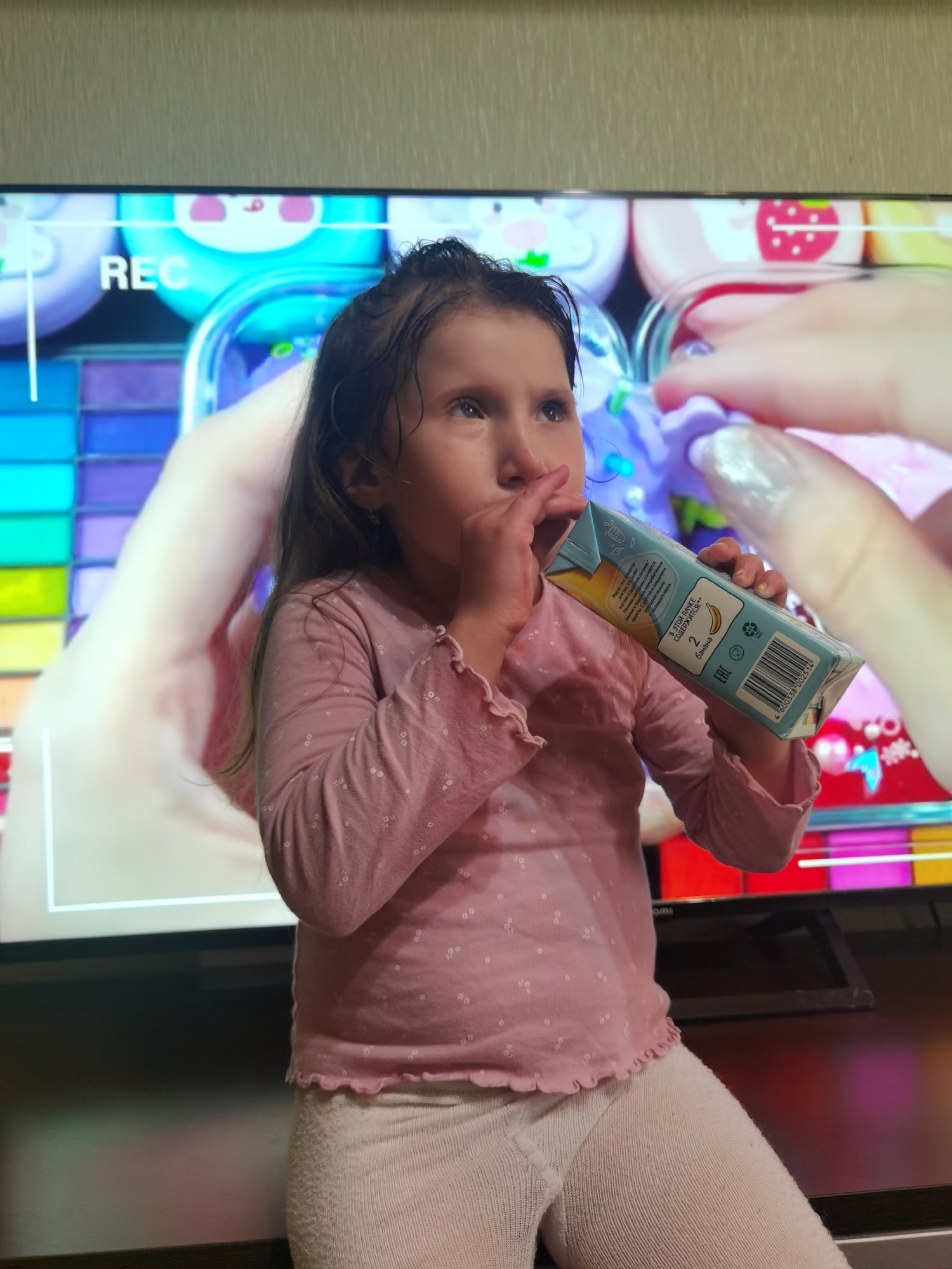 A young child, appearing to be a girl with dark, slightly damp hair, is seated on a dark, flat surface in front of a large flat-screen television. She is wearing a light pink long-sleeved top with a white polka-dot or floral pattern and light-colored, possibly off-white, ribbed leggings. She holds a small, light blue juice carton in her left hand, with a straw inserted, and is drinking from it, looking upward and to the right. The carton's label, written in Cyrillic script, identifies the contents as banana, with the text "В ЭТОЙ ПАККЕ СОДЕРЖИТСЯ... 2 банана" (This pack contains... 2 bananas) and the brand "ФрутоНяня" partially visible. A barcode (460338806214) and recycling symbols are also present on the carton.
The television screen displays vibrant animated content featuring various cartoon elements, including a pink pig and a red strawberry icon. The text "REC" in white sans-serif font is visible in the upper left corner of the screen, suggesting a recording interface or content related to recording. A bare hand with silver glitter nail polish on the fingernails is visible on the right side of the screen, seemingly interacting with a purple object depicted in the on-screen content. The immediate background above the television consists of a light-colored wall with a subtle textured pattern. The image provides no visual information to confirm the location in the city of Chaikovsky, Russia, beyond the Russian text on the juice carton indicating a product from Russia.