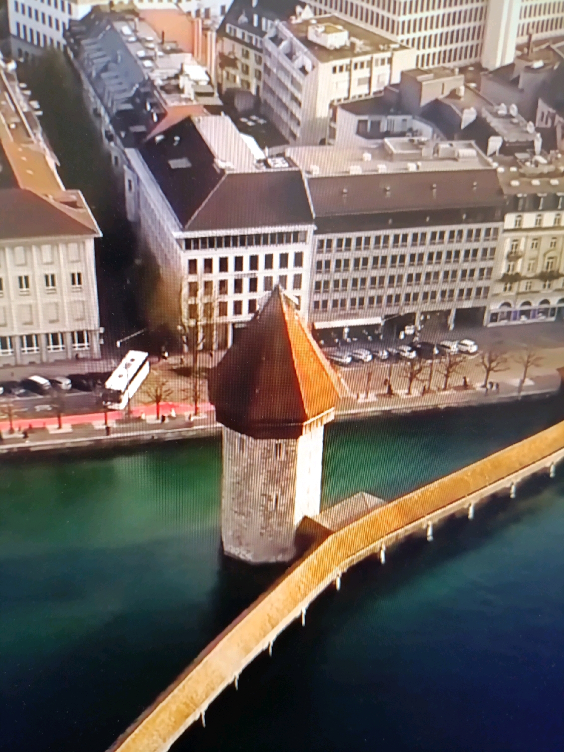 The image depicts an aerial view of the Chapel Bridge (Kapellbrücke) and the Water Tower (Wasserturm) in Lucerne, Switzerland, not Netanya, Israel. The Chapel Bridge is a covered wooden footbridge spanning the Reuss River, adorned with triangular roof tiles. The Water Tower, a cylindrical stone structure with a conical roof, stands adjacent to the bridge. The river's water is a deep green hue. In the background, several multi-story buildings with traditional European architecture line the riverbank, some featuring pitched roofs and multiple windows. A white bus is visible on the street beside one of the buildings, along with parked cars. The scene is characterized by a clear day with ample sunlight, casting shadows on the buildings and water.