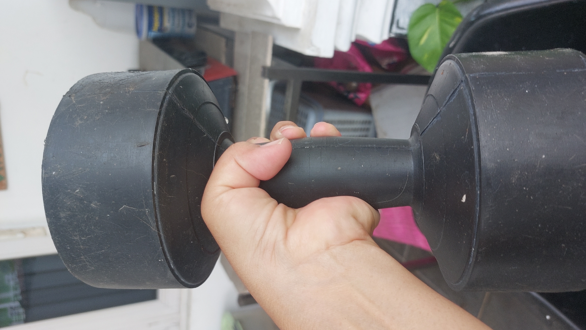 A close-up, eye-level, outdoor shot captures a person's right hand firmly gripping the black, rubberized handle of a dumbbell. The hand is flesh-toned and appears to be in mid-grip, with fingers wrapped around the cylindrical middle section of the weight. The dumbbell itself is substantial, with large, circular weights at either end, both covered in a layer of dust and fine scratches, indicating it is well-used.
The background is blurred, suggesting a shallow depth of field, but hints at an outdoor environment, possibly a porch or yard in Bandar Setia, Indonesia. To the left, a white-framed window with vertical blinds is visible, with faint greenery beyond. To the right, several out-of-focus objects are present, including what appears to be a blue and yellow container, a dark table with a dark object on it, and a potted plant with large, green leaves. The overall impression is one of a casual fitness activity taking place in a residential setting.