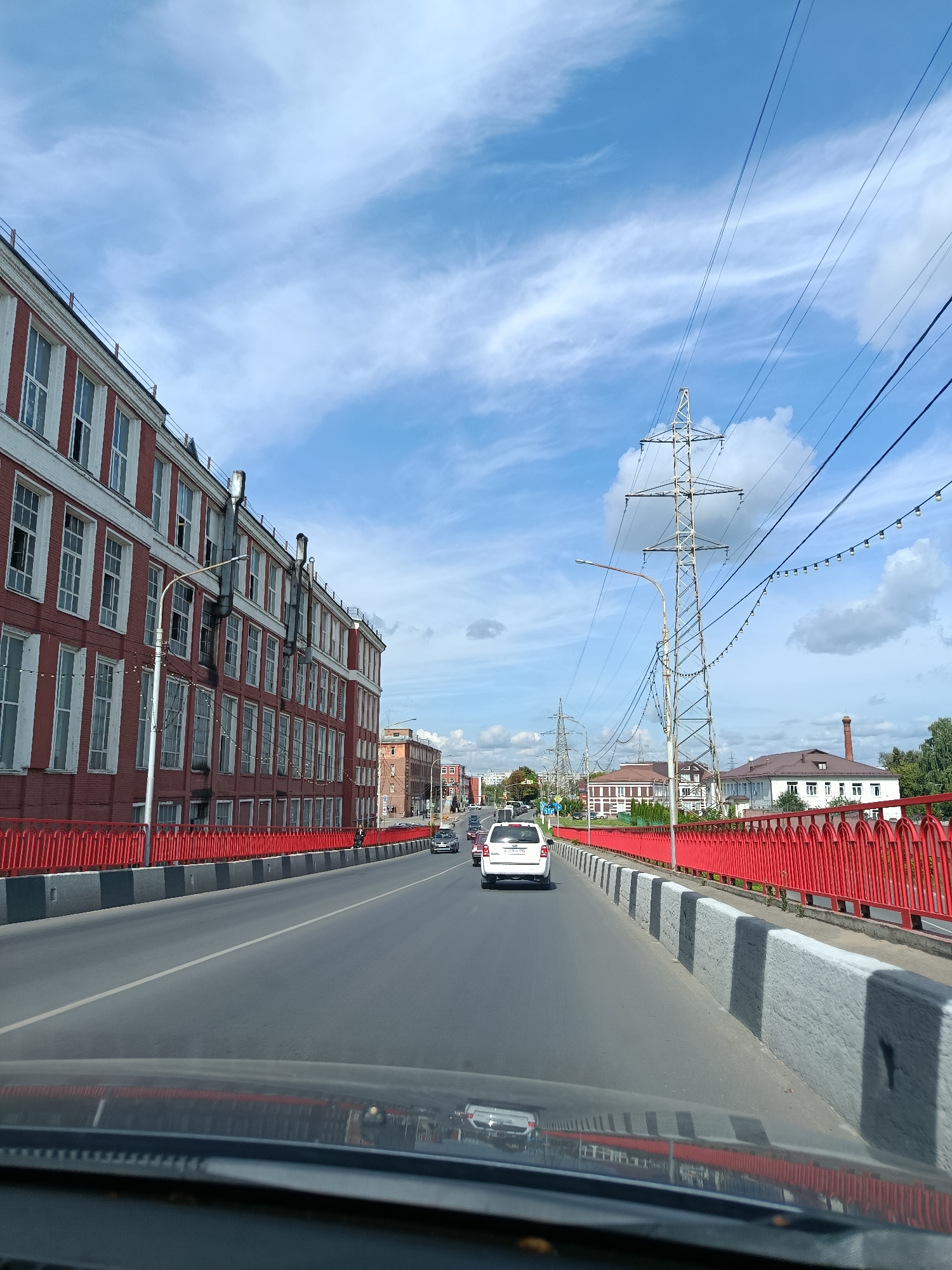 The image depicts a view from inside a vehicle traveling on a bridge in Orekhovo-Zuyevo, Russia. The bridge features a bright red railing along its sides. To the left of the road is a large, multi-story brick building with numerous white-framed windows. Beyond this building, further down the road, other structures can be seen, including a tall brick chimney. On the right side of the bridge, there are also buildings and vegetation. Overhead, power lines are strung between tall pylons. Several vehicles are visible on the road, including a white SUV directly ahead and other cars further in the distance. The sky is blue with scattered white clouds. The scene appears to be captured during daylight hours, likely late morning or early afternoon, based on the lighting conditions.