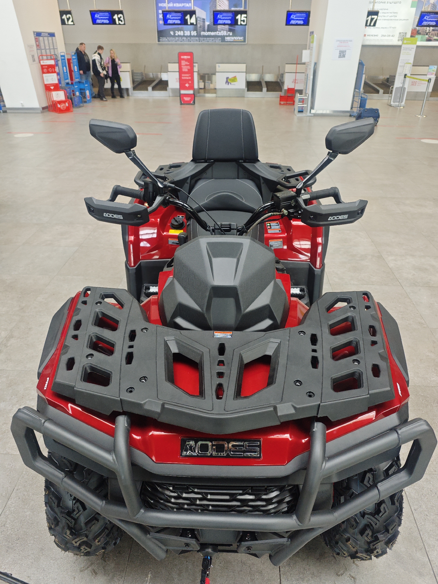 The image shows a red AODES brand ATV (all-terrain vehicle) parked inside what appears to be the Bolshoye Savino airport terminal in Perm, Russia. The ATV is the primary subject, positioned centrally in the foreground. It features black front and rear cargo racks, hand guards labeled "AODES," and heavy-duty tires.
In the background, airport features such as check-in counters with electronic signs displaying flight information (likely destination “Perm”), luggage conveyor belts, and queue dividers are visible. People are standing near check-in counters 12 and 13. A display of promotional materials with "НОВЫЙ КВАРТАЛ" is seen on a sign above counter 14, along with contact information "248 38 95 moments59.ru."
The environment is an airport terminal, characterized by a clean, tiled floor and bright lighting.