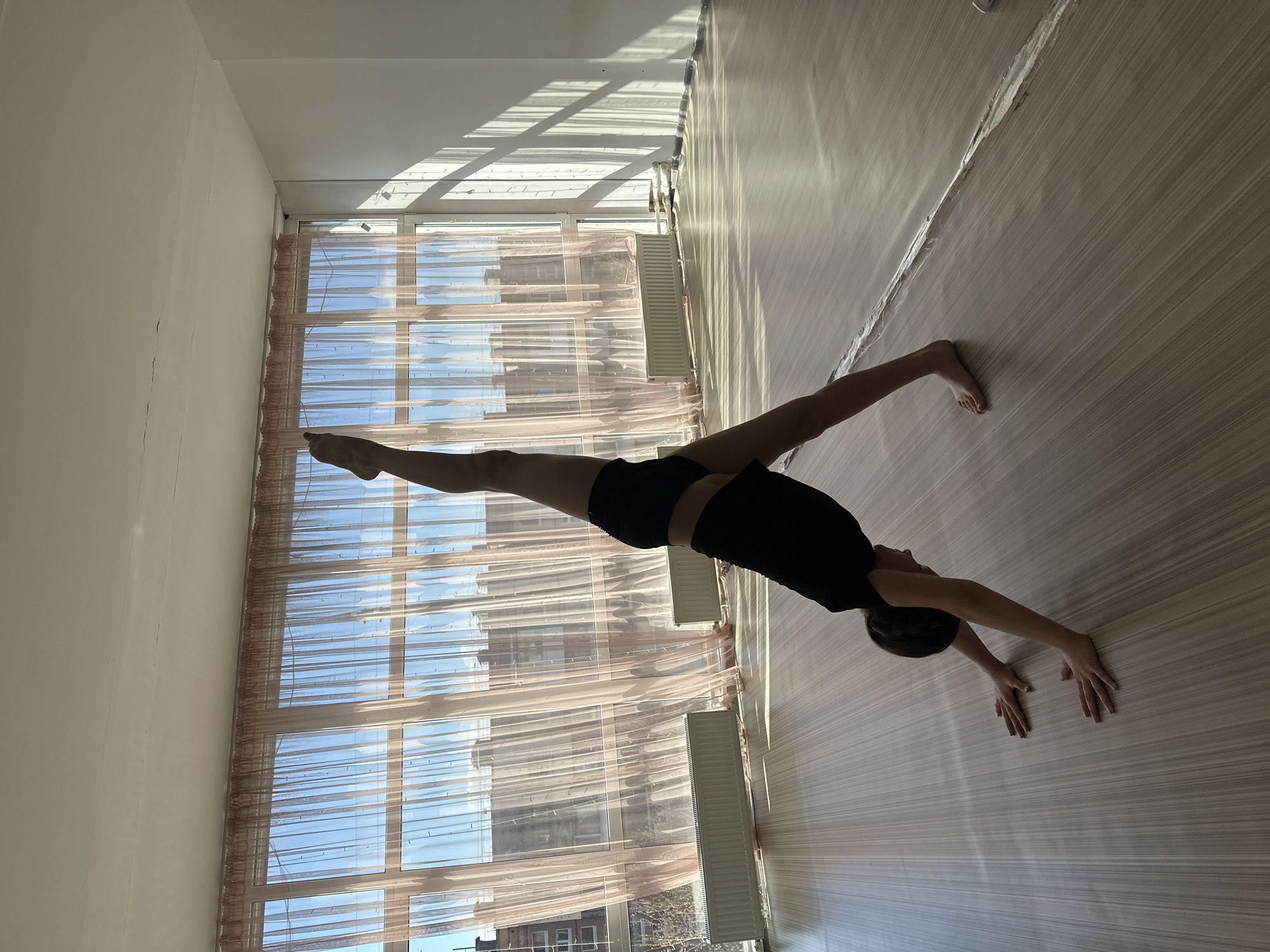 The image, rotated 90 degrees clockwise, displays an indoor scene where a person is engaged in a physical activity. A young individual, with dark, short hair and clad in a black t-shirt and black shorts, is performing a gymnastic or yoga pose, resembling a deep backbend or bridge pose. They are balancing on their hands and feet, with their body arched towards the ceiling and their face partially visible, looking upwards.
The room features a light-colored wooden or laminate floor with noticeable parallel grain patterns. The walls are plain and off-white, with some faint cracks visible on the wall to the left. A large window with multiple panes covers a significant portion of one wall, adorned with sheer, light-colored (possibly a delicate pink or beige) curtains. Through the window, a bright blue sky is visible, along with vague outlines of buildings in the distance, indicating an outdoor view.
Strong sunlight streams through the window, casting sharp, elongated shadows of the window frames onto the floor and wall, suggesting a clear day, likely during the morning or afternoon. Two white radiators are positioned below the window, implying a need for heating. Some pipes and fixtures are also visible near the top of the window area. No discernible text is visible anywhere in the image. The scene portrays a moment of focused physical exertion within a well-lit, functional interior. This location is identified as Votkinsk, Russia.