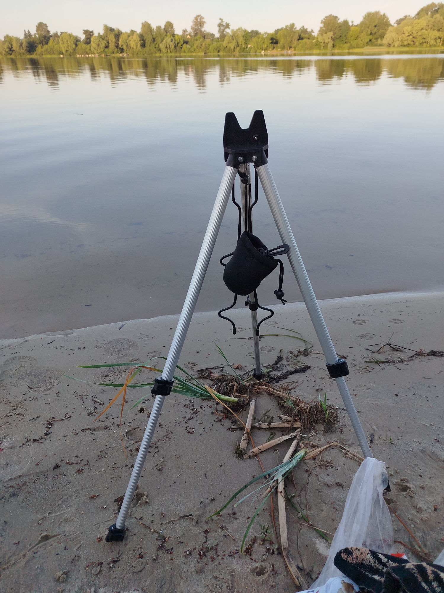 The image shows a tripod on a sandy beach next to a body of water. The tripod is silver with black accents and has a small black pouch attached to it. There are some dry reeds and other debris scattered on the sand around the tripod. In the background, there is a calm body of water with reflections of trees and the sky. The lighting suggests it is either early morning or late afternoon. The scene appears to be in a natural environment, possibly a park or a natural reserve.
