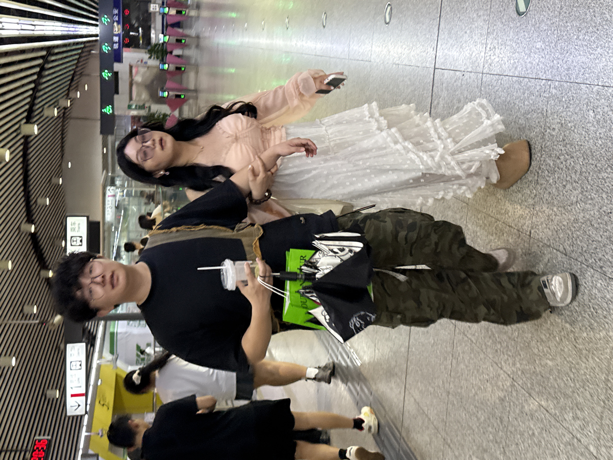 The photograph depicts two individuals in what appears to be an indoor public space, likely a transportation hub or commercial complex.
The primary subject is a young man wearing glasses, a black t-shirt, and camouflage pants. He is holding a cup with a straw in one hand and an umbrella and a green shopping bag in the other.
Beside him stands a woman with long black hair wearing glasses, a light peach-colored top, and a white, lace-detailed dress. She is holding a phone in her hand.
The background reveals a modern interior with a tiled floor and a ceiling with linear light fixtures. In the distance, there are signs with symbols and Chinese characters. Other individuals are walking nearby.
Given the presence of Chinese characters on signage and architectural styles, the location is most likely in China, consistent with the city mentioned. The specific location within Yuzhong District cannot be determined based on the image alone.