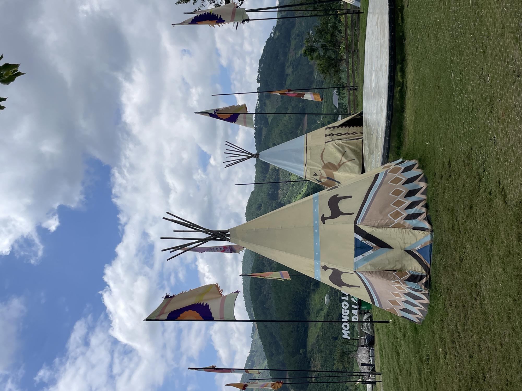 This outdoor image captures a themed recreational area in Thôn 5, Vietnam, under a bright, partly cloudy blue sky. The foreground is dominated by neatly mown green grass. Three teepee-style tents are visible, crafted from tan fabric. The largest tent, positioned in the left foreground, features elaborate geometric patterns in blue, brown, and white near its base, topped by a light blue band and two dark brown silhouettes of llamas or alpacas. A second tent, located behind and to the right of the first, displays a darker silhouette of a human figure riding an animal, possibly a deer, alongside geometric patterns at its base. A third tent is partially visible on the far right edge of the frame.
Numerous decorative flags on tall black poles are scattered across the scene. Several flags feature stylized sun-like designs in colors such as orange, purple, blue, and green, appearing somewhat frayed at their edges. A large, light-colored circular platform with a dark rim is situated on the grass in the mid-ground, to the right of the central teepee. In the distant left mid-ground, a white sign with dark lettering, partially reading "MONGOL LA DAL," is visible next to a few small, low-lying structures with thatched or tarp coverings. The background consists of a continuous range of green, forested hills under the expansive sky. No individuals are present in the image.