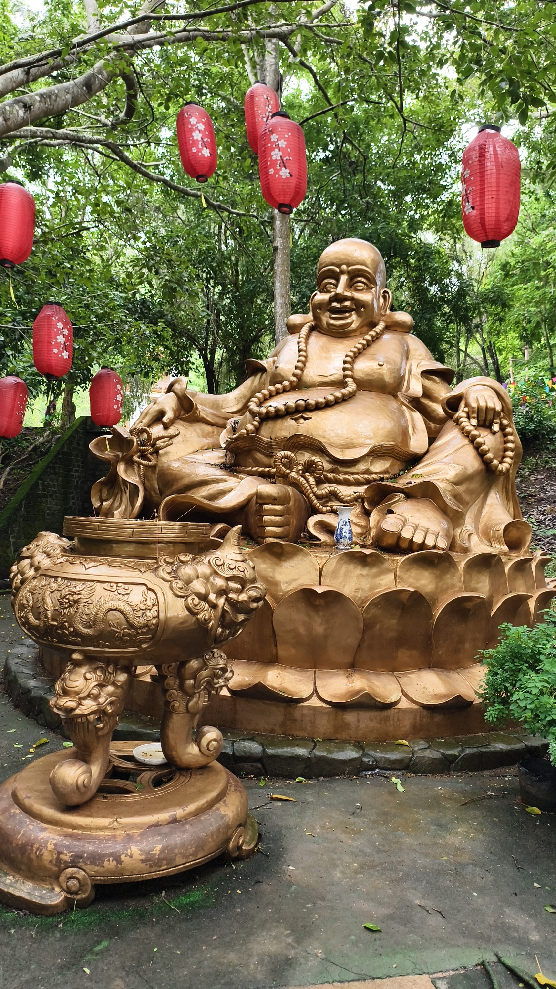 The image shows a large, golden statue of a laughing Buddha sitting on a lotus flower base in an outdoor setting. The statue is adorned with a beaded necklace and is positioned in front of a golden incense burner supported by decorative legs. Red paper lanterns with floral designs are suspended from the trees in the background. The ground is paved and wet, with fallen leaves scattered about. Lush green foliage surrounds the statue and forms a dense canopy overhead. The scene appears to be in a temple or garden, possibly located in Tân Phú, Vietnam, given the context of the location provided.