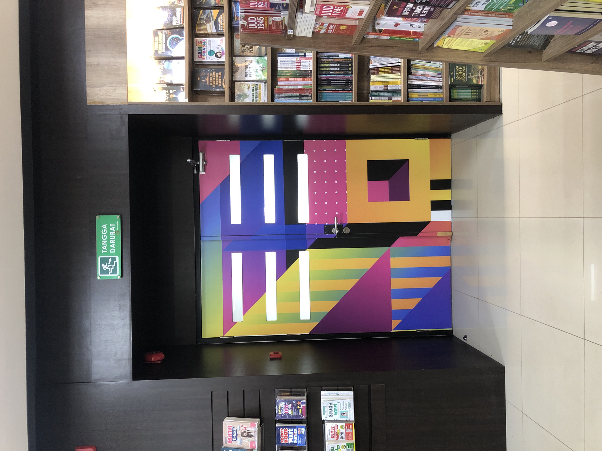 The image shows the entrance to a bookstore, likely a Gramedia, in Banda Aceh, Indonesia, suggested by the "Gramedia" text on the emergency exit sign. The main subject is a brightly colored, abstractly designed double door, featuring geometric shapes in shades of purple, pink, yellow, blue, green, and orange. Above the door, an illuminated green emergency exit sign with the text "TANGGA DARURAT" (Emergency Stairs) is visible.
To the left of the door, a dark brown wooden cabinet with multiple shelves displays various books and educational materials. Some of the visible book titles include "MATH TRICKS," "POCKET BOOK MATE MATIKA," "MATH IPA," "Study TRICKS," and "RUMUS MATEMATIKA SD/MI."
To the right of the door, a wooden bookshelf is filled with a large collection of books. Titles and spines suggest a variety of genres, including educational materials and legal texts like "UUD 1945" (The 1945 Constitution of Indonesia). The flooring is a light-colored tile. The overall environment appears to be a well-lit indoor retail space.