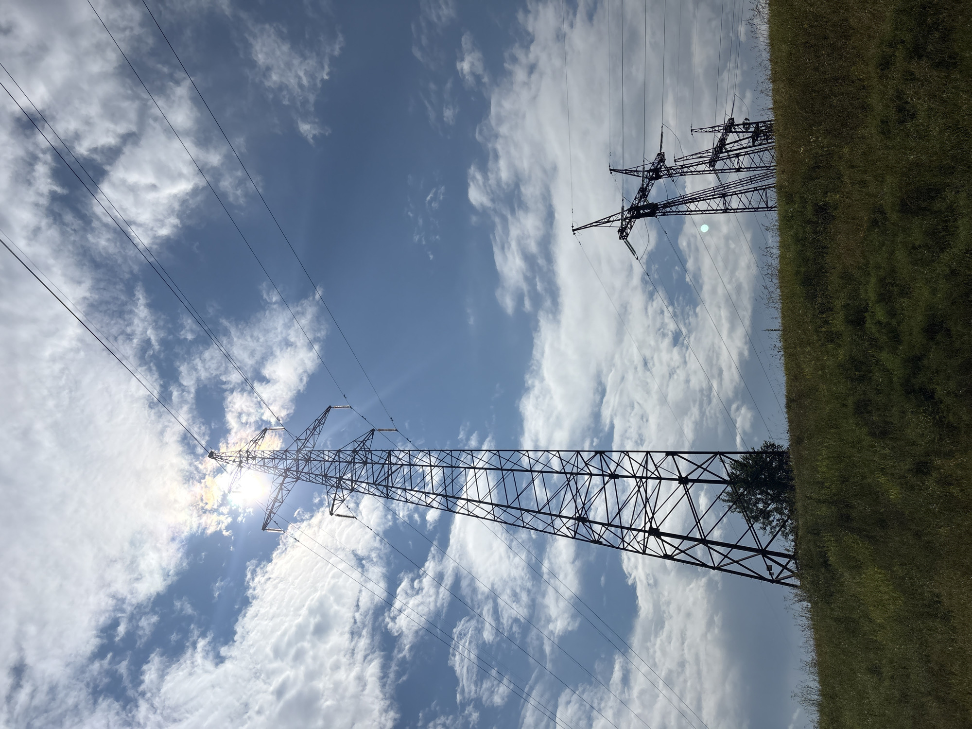 This is an eye-level, medium shot photograph of power line towers against a partly cloudy sky. The sun is partially obscured by clouds, creating a bright glare. In the foreground, a grassy embankment slopes upwards. Three metal power line towers are visible, with the tallest one on the left. The towers are silhouetted against the bright sky and are connected by thick, dark wires. Some trees and vegetation are visible on the embankment near the base of the towers. The image does not depict any people. The location is unknown.