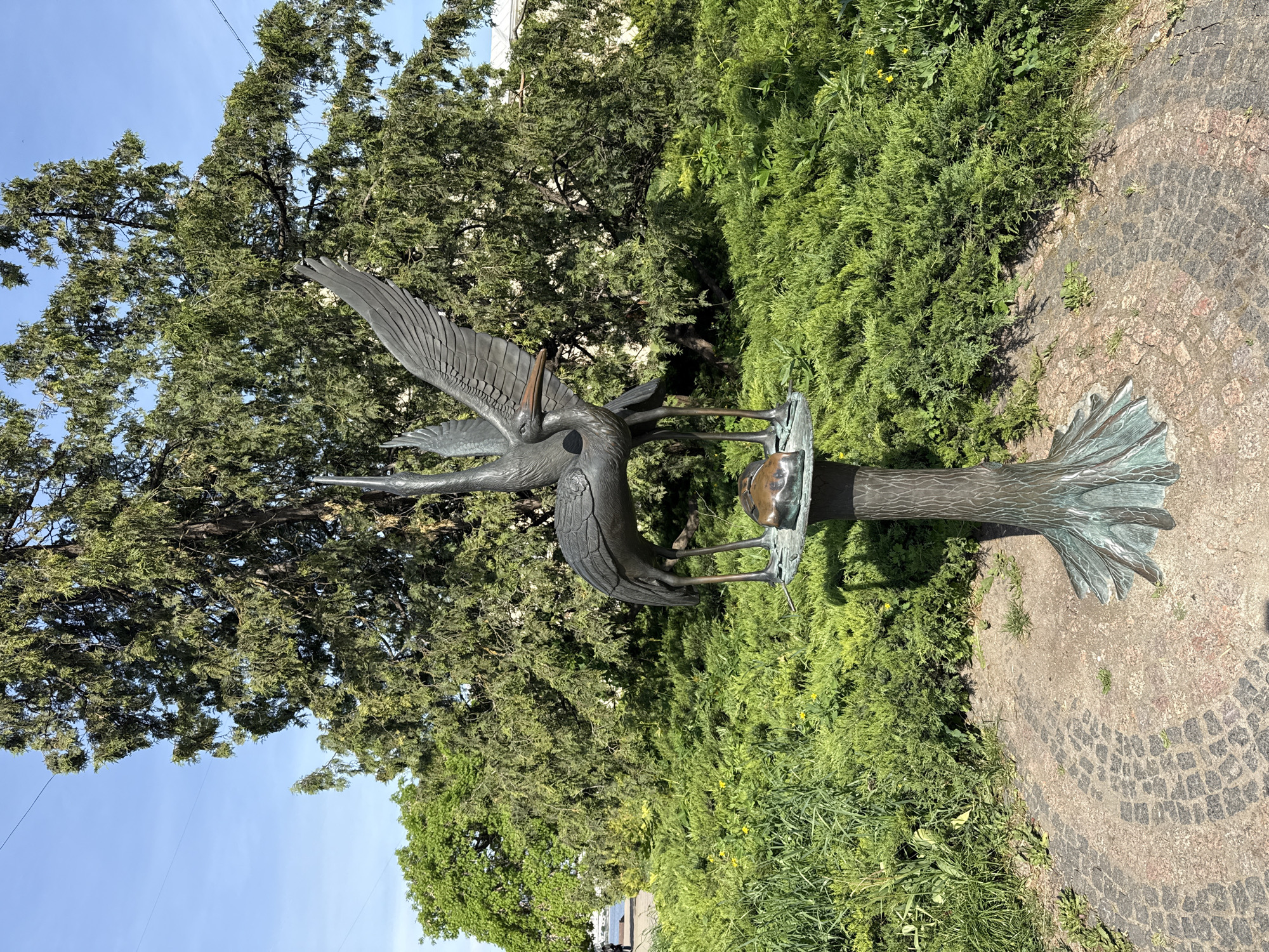The image captures a bronze sculpture located in the city of Ternopil, Ukraine. The sculpture depicts two herons perched around a bird feeder. The sculpture is mounted on a tree stump made of the same bronze material. The surrounding environment includes lush green bushes and a large tree in the background. The ground around the sculpture is paved with cobblestones. The sky is visible in the upper left corner and is a clear, bright blue.